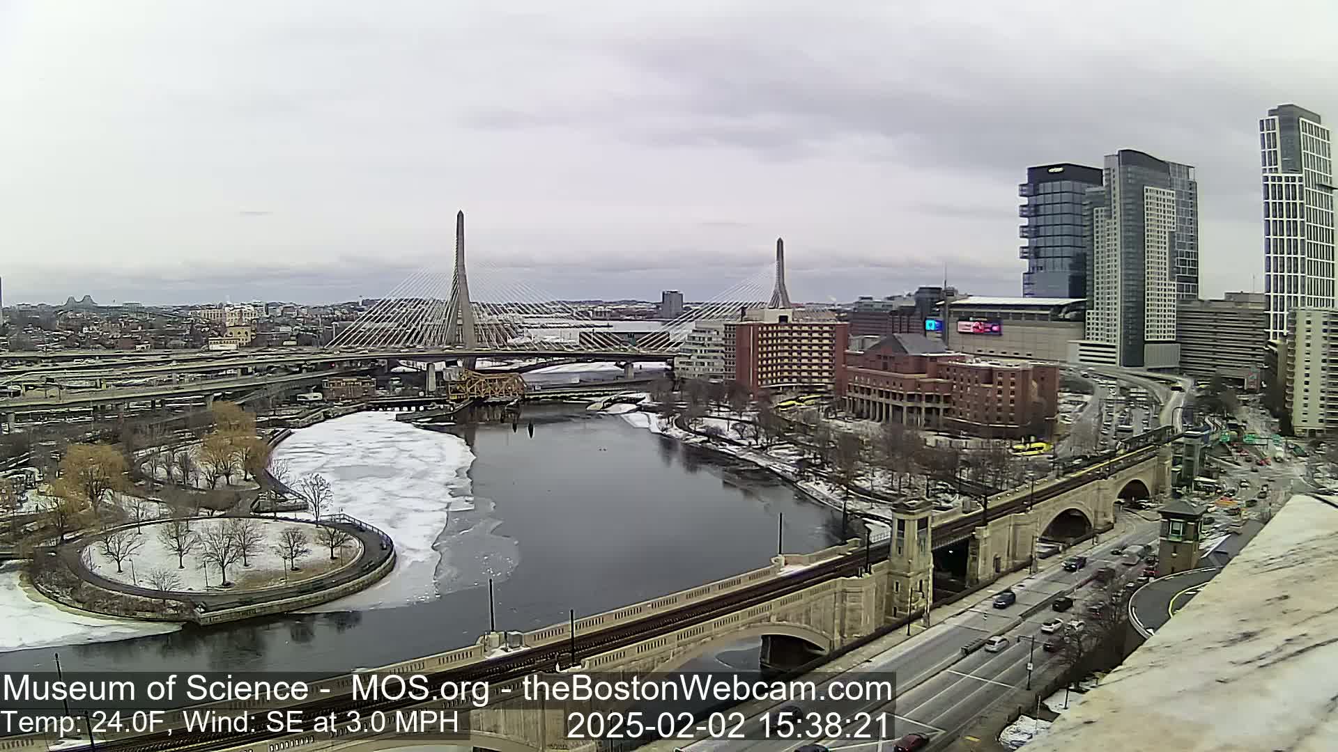 A city scene on an overcast day shows a river partially covered in ice, two cable-stayed bridges, and a cityscape with modern high-rises.