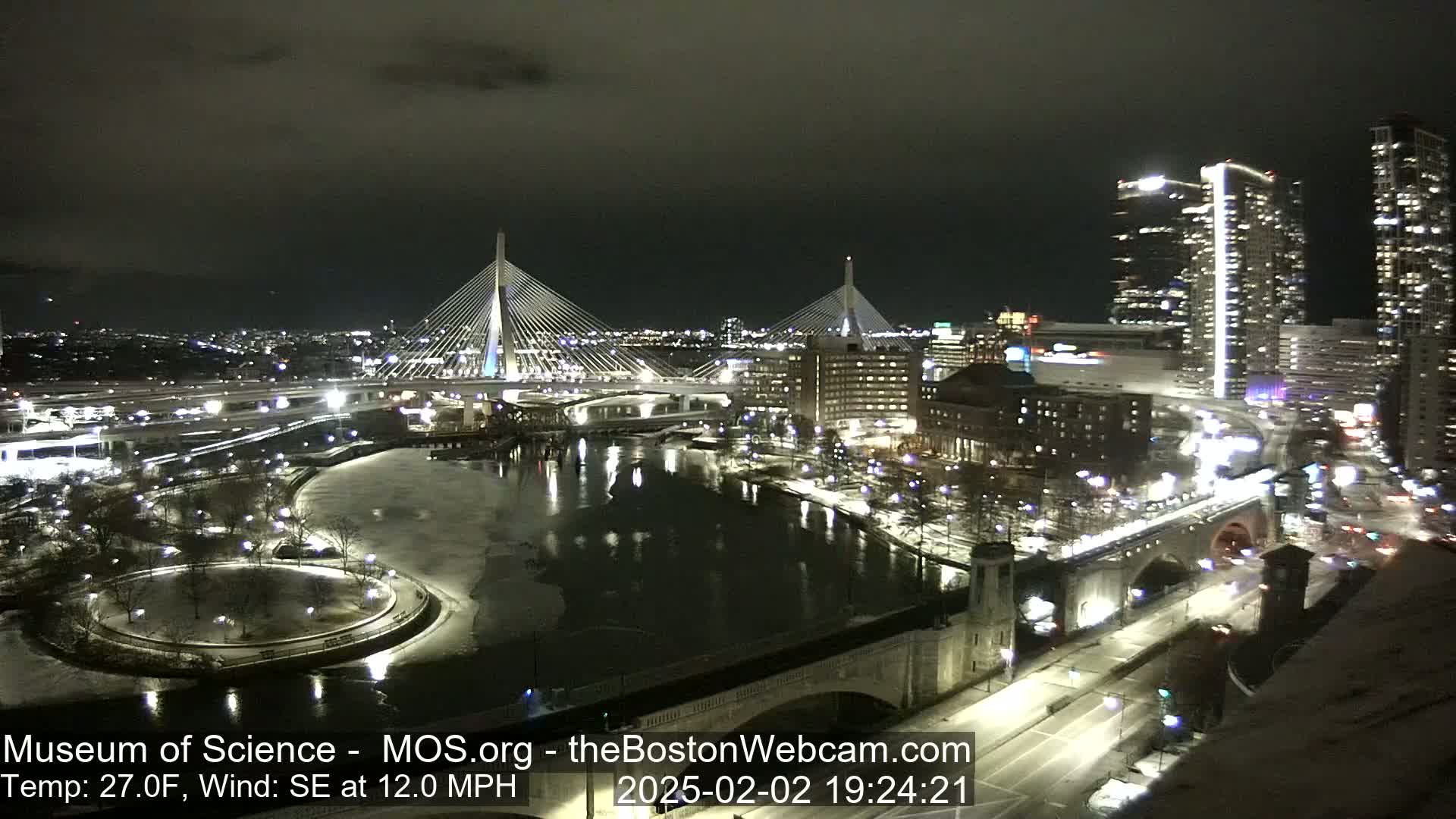 A nighttime city scene shows two cable-stayed bridges spanning a partially frozen river, with snow-covered ground and city lights illuminating the area under a dark sky, with a southeast wind.