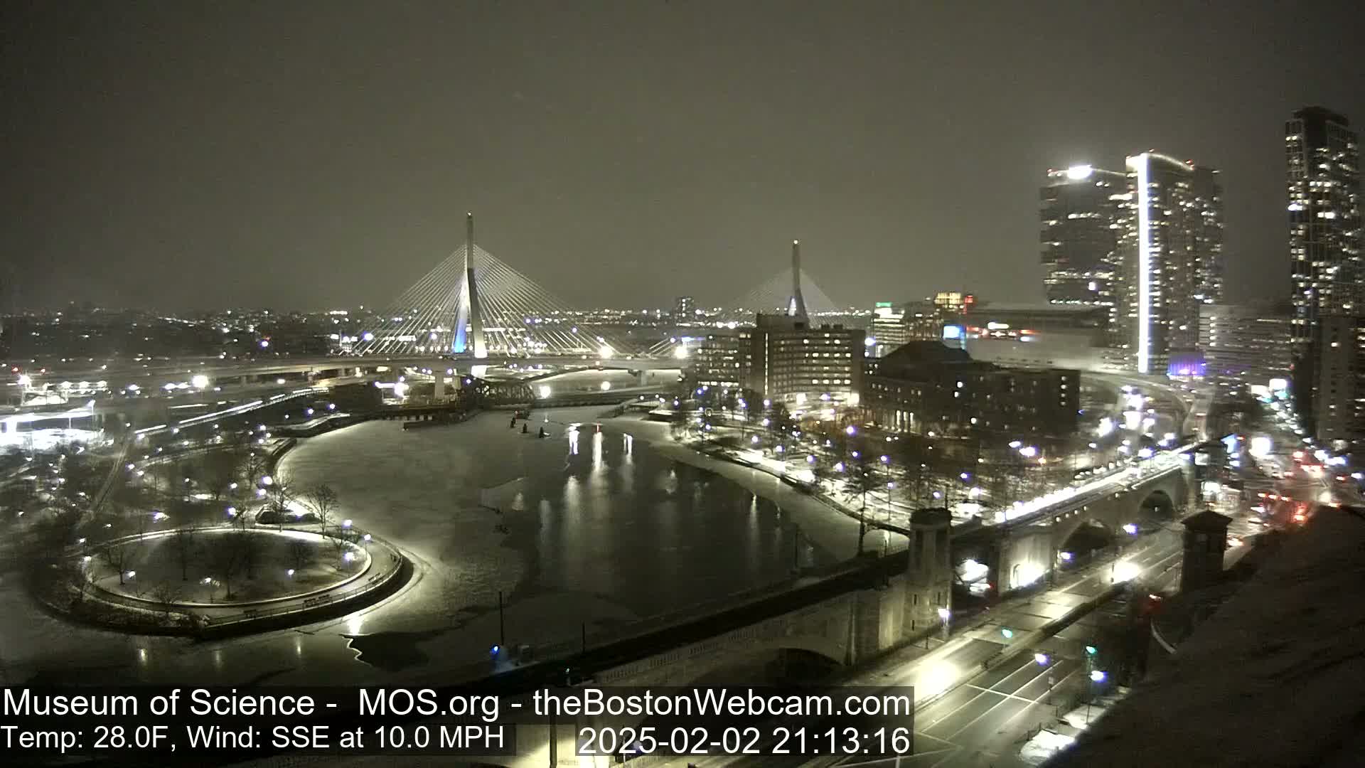 A nighttime, snowy cityscape features two illuminated cable-stayed bridges spanning a partially frozen river, with city lights reflecting on the water's surface under a dark sky.
