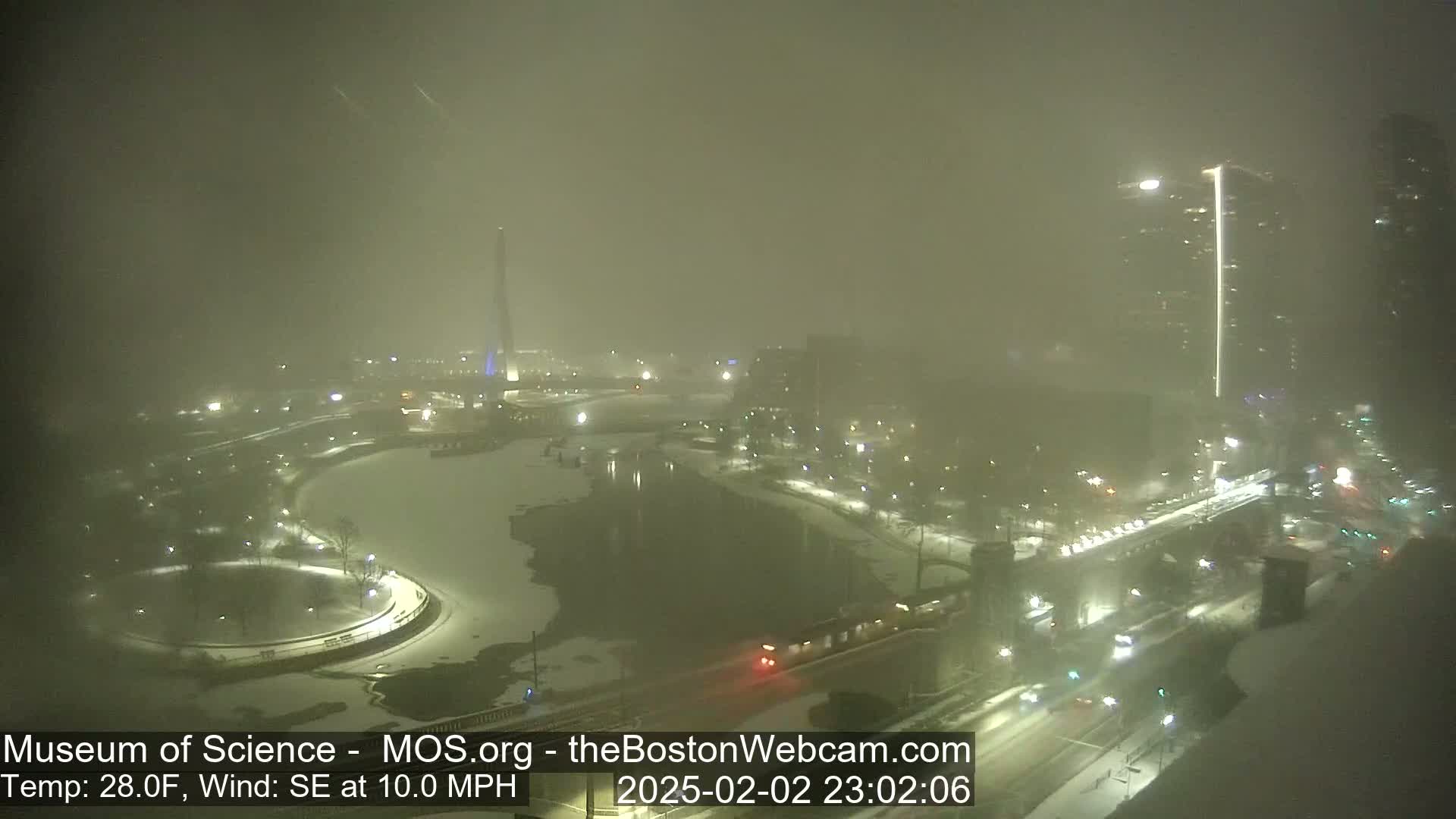 A snowy, foggy nighttime scene shows a city with a river, a monument, and buildings, with a southeast wind at 10 mph and a temperature of 28 degrees Fahrenheit.
