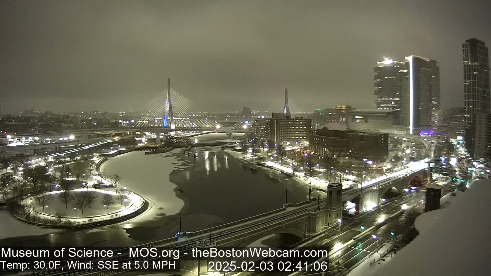 A snowy nighttime cityscape shows two bridges, a river partially frozen over, and city buildings illuminated under a gray sky.