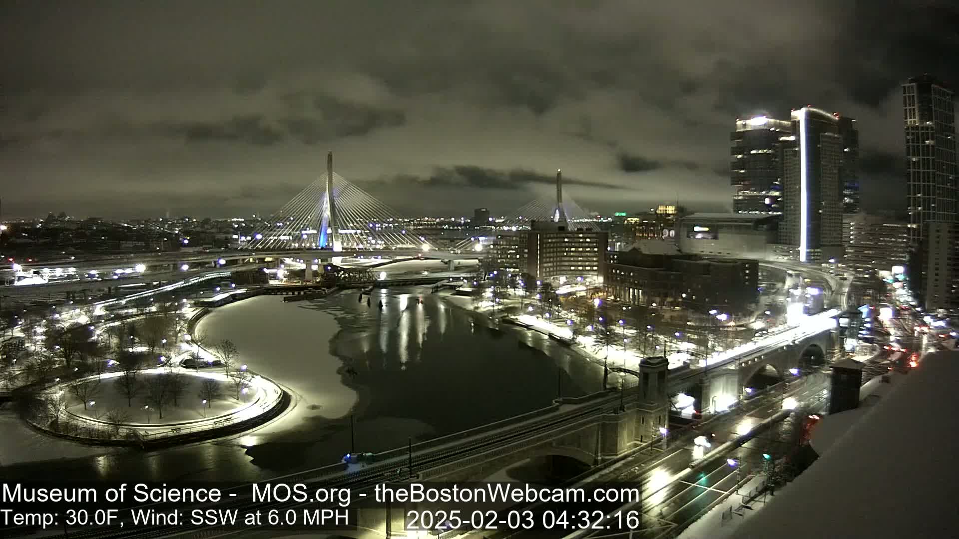 A nighttime view of a city with snow-covered ground and a partially frozen river, illuminated bridges, and tall buildings under a cloudy sky.