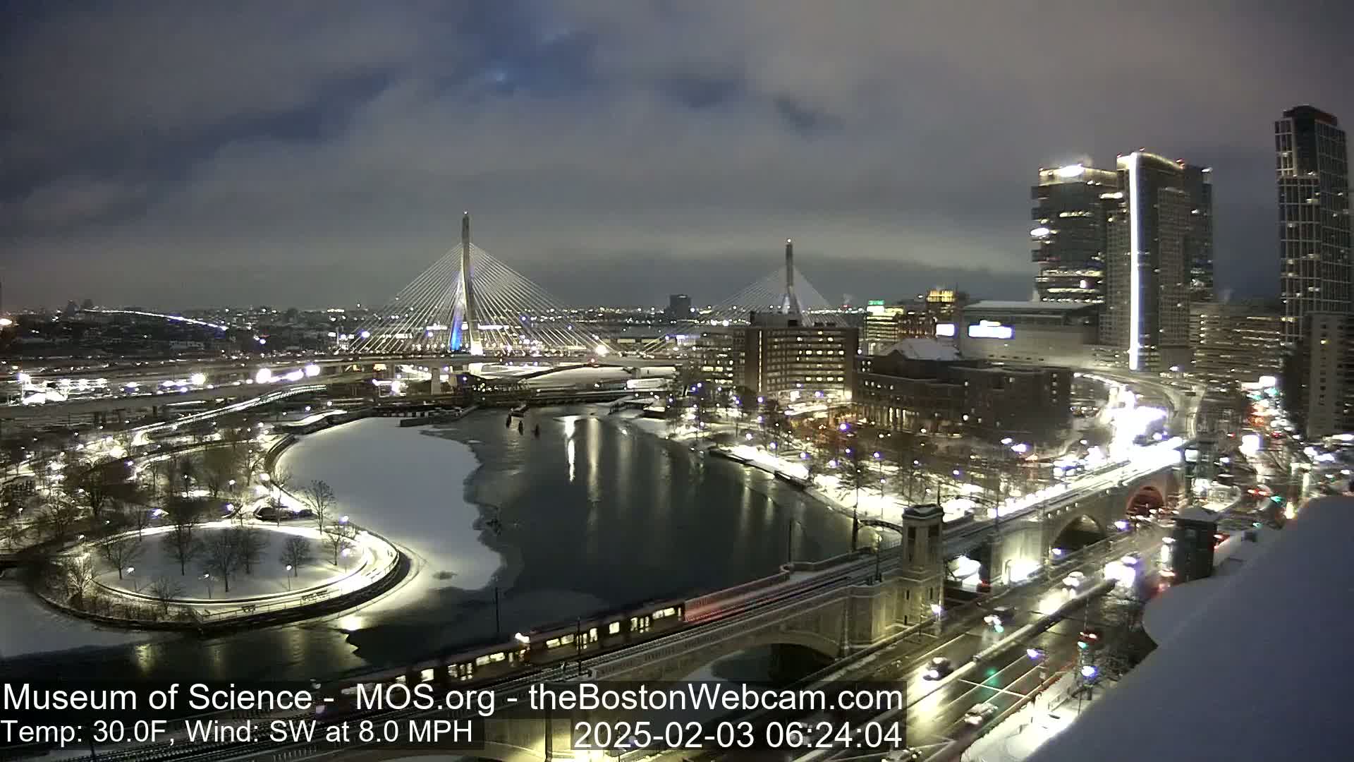 A nighttime cityscape with snow-covered ground, partially frozen water, and two cable-stayed bridges under a cloudy sky.