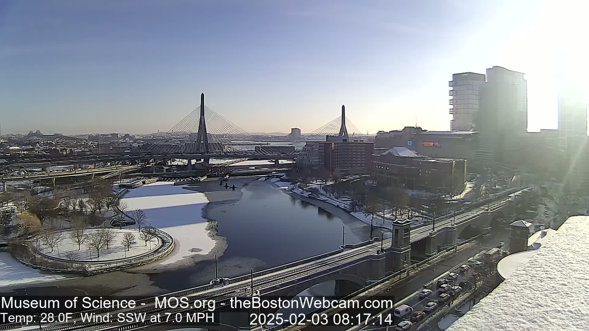 A sunny, snowy cityscape featuring two cable-stayed bridges spanning a river, with partially frozen water and a snow-covered park.