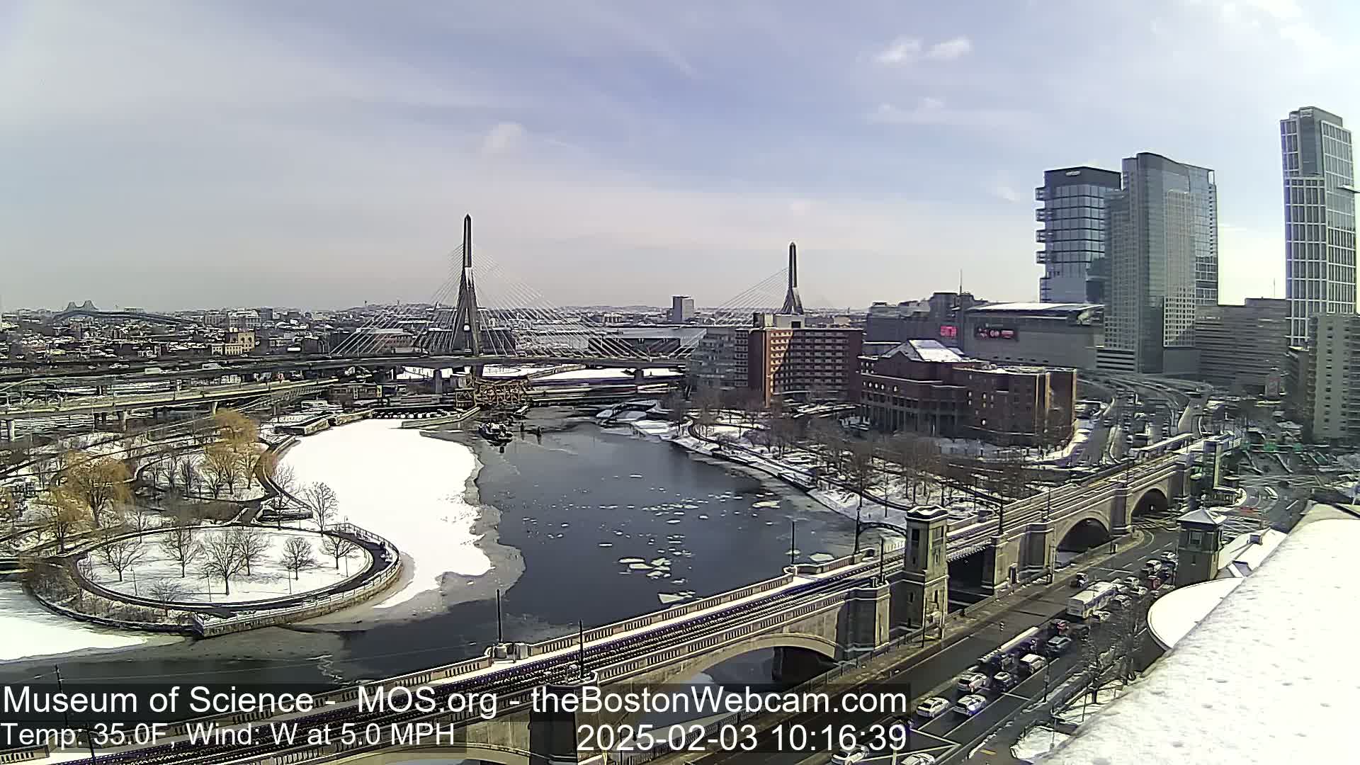 A partly sunny winter day reveals a cityscape with bridges, a river partially covered in ice, and snow-covered ground.