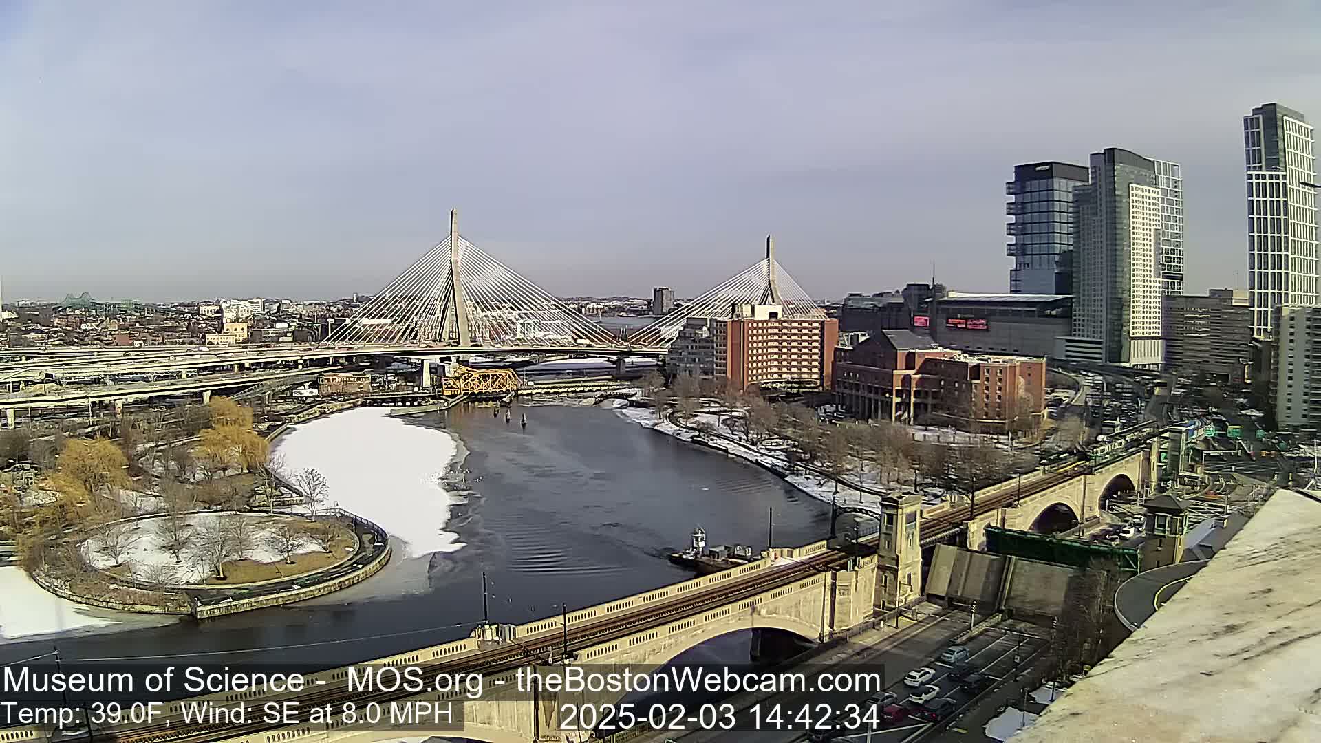 A partly sunny day reveals a cityscape with two cable-stayed bridges spanning a river partially covered in ice, alongside other bridges, roads, and buildings; some areas show patches of snow.