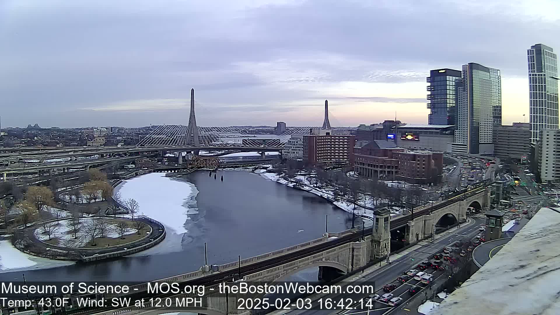 A partly frozen river flows beneath two cable-stayed bridges, passing through a city with modern high-rises and a partially snow-covered landscape under a cloudy sky.