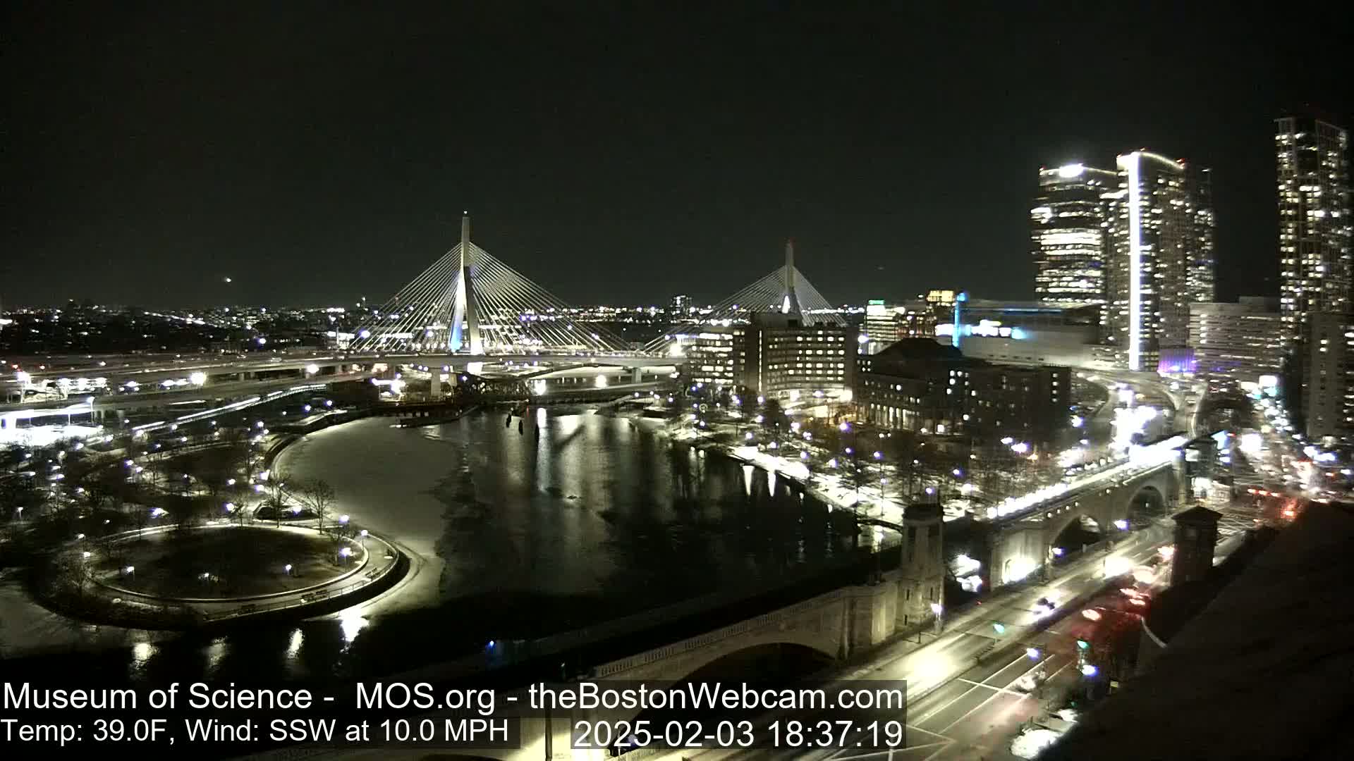 A nighttime cityscape featuring two cable-stayed bridges, a river with some ice, and city buildings illuminated, under a clear sky with a 39°F temperature and a 10 mph south-southwest wind.
