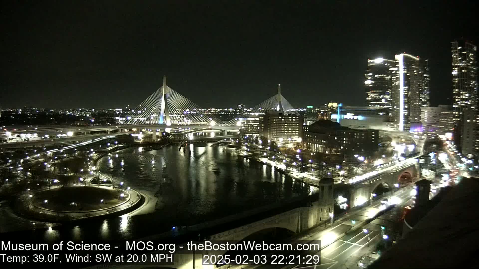 A nighttime city scene shows two illuminated cable-stayed bridges spanning a dark river, with city lights reflecting on the water, under a clear sky and a southwest wind of 20 mph.