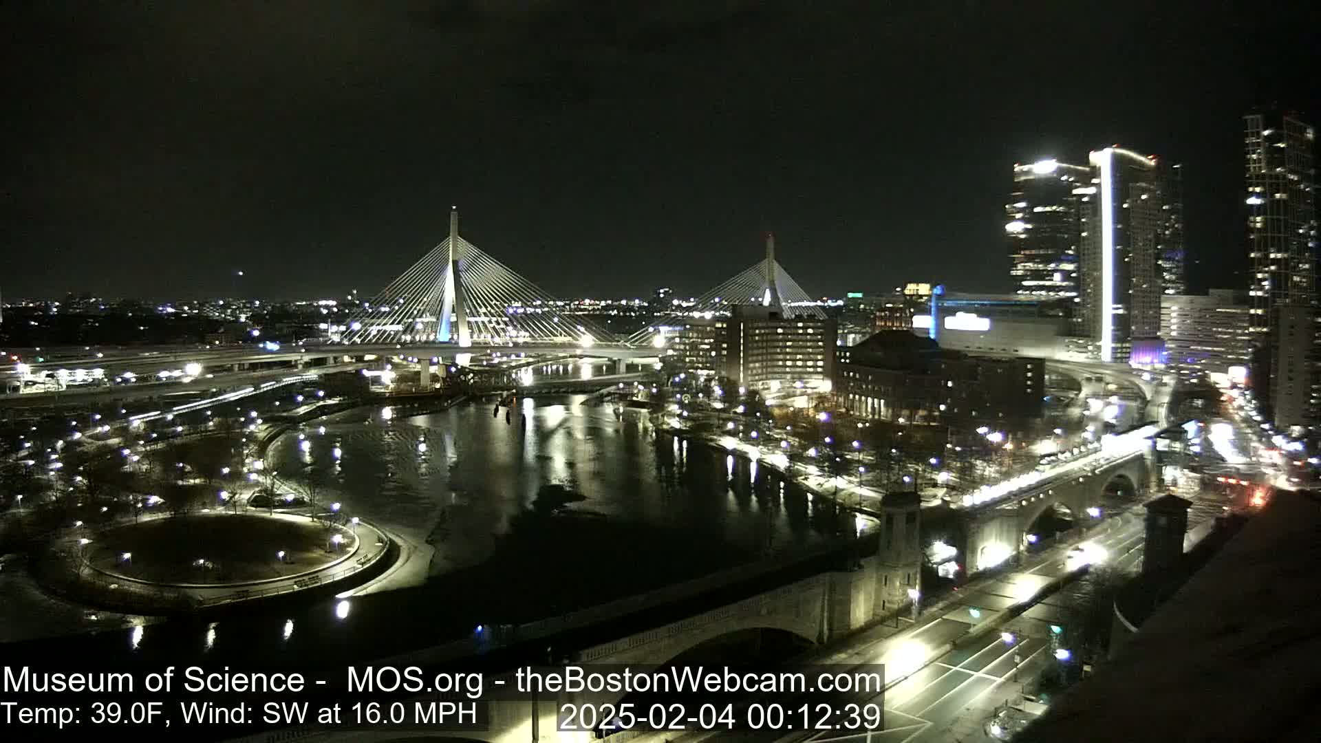 A nighttime view of a city waterfront with two illuminated cable-stayed bridges, a river, and city buildings, under a clear, dark sky with a southwest wind of 16 mph and a temperature of 39°F.