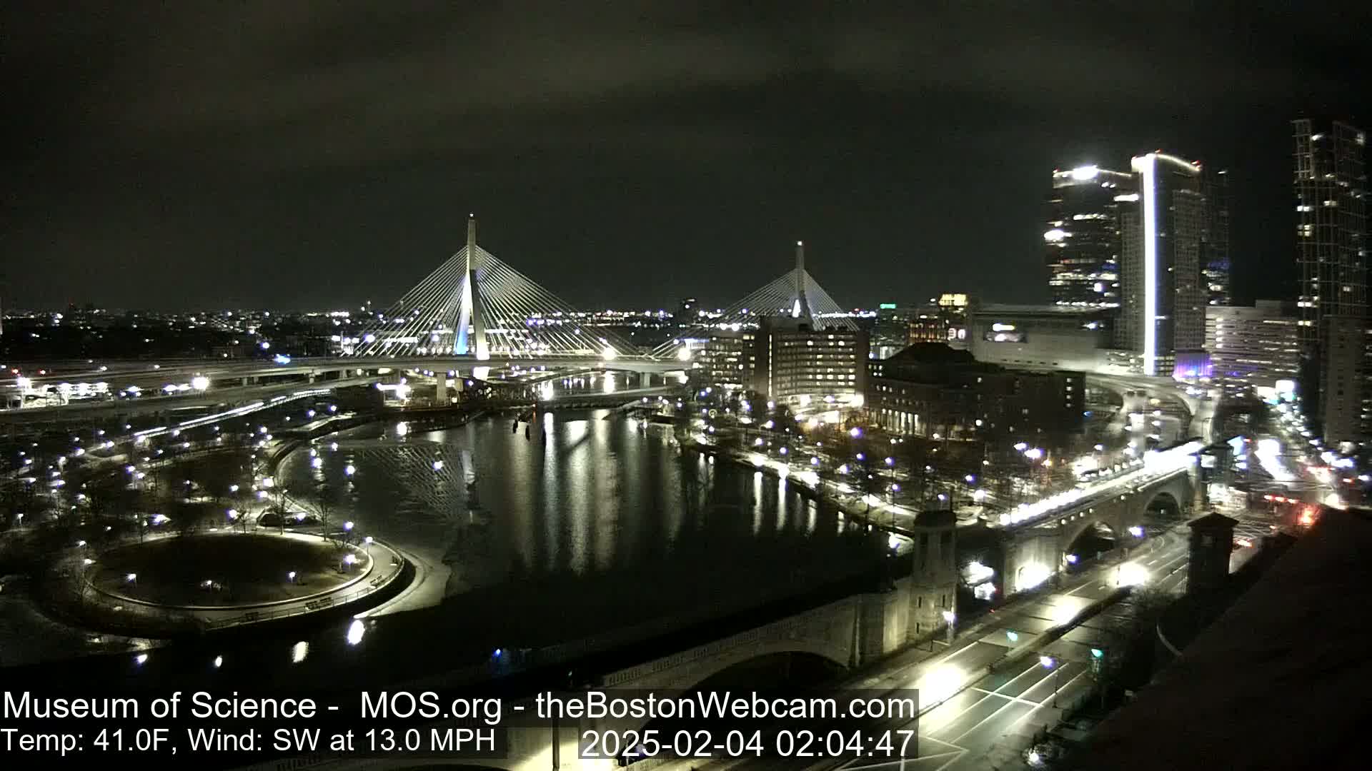 A nighttime city scene shows two illuminated cable-stayed bridges spanning a dark river, with city lights and buildings reflecting on the water, under a dark, clear sky and a southwest wind.