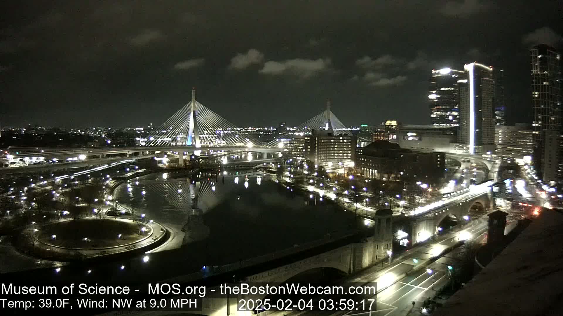 A nighttime cityscape shows two illuminated cable-stayed bridges spanning a river, with city lights reflecting on partially frozen water under a partly cloudy sky.