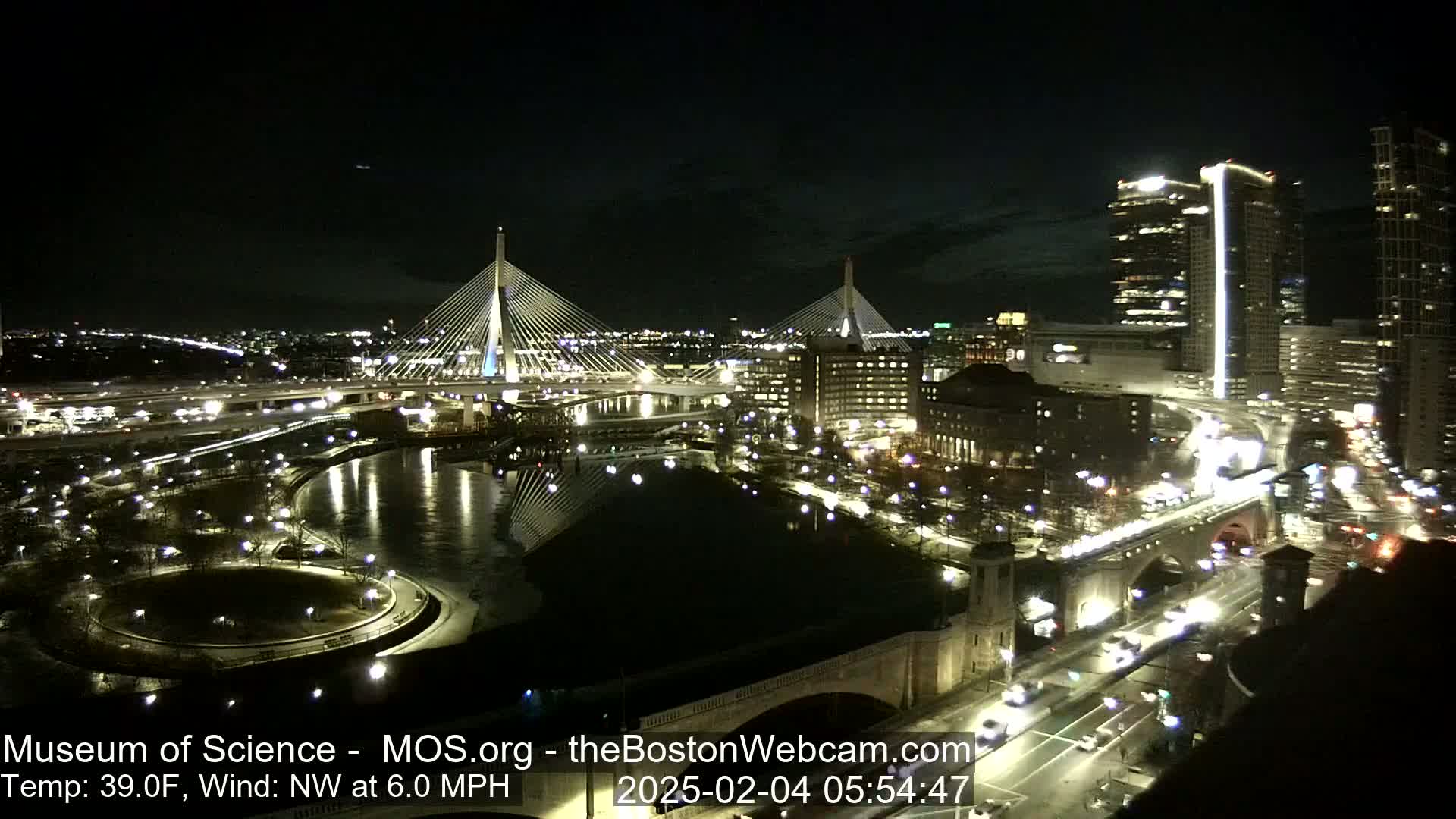 A nighttime aerial view of a city with two cable-stayed bridges, a river, and roadways, under a clear, cool sky with a northwest wind.
