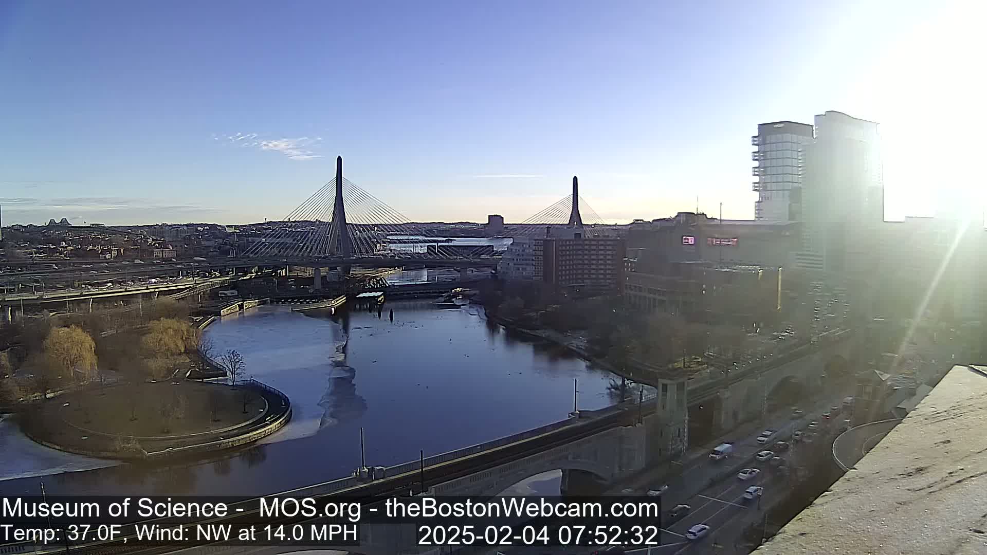 A partly sunny day with a northwest wind of 14 mph and a temperature of 37°F shows a city view of two cable-stayed bridges spanning a river with partially frozen water, surrounded by buildings and roads with cars.