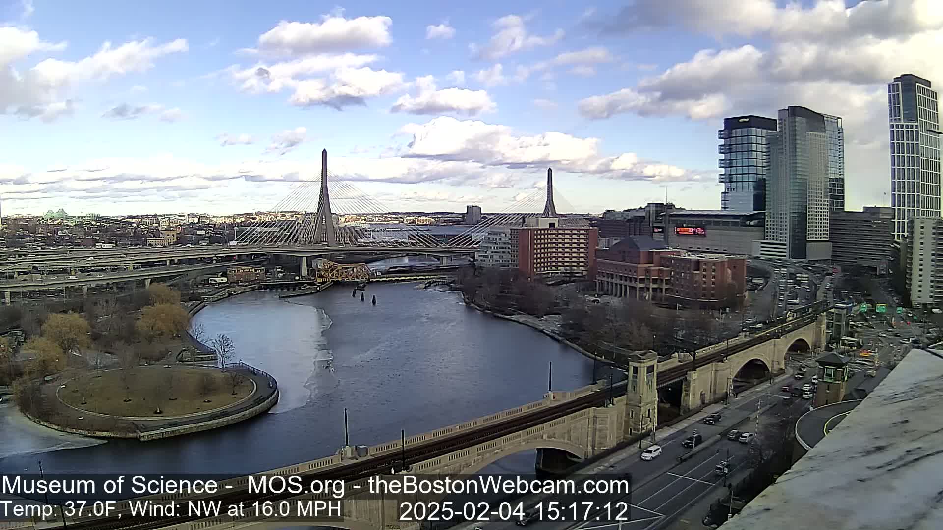 A partly cloudy day shows a city with two cable-stayed bridges crossing a river, partially frozen, with a city skyline, including several tall buildings, in the background.