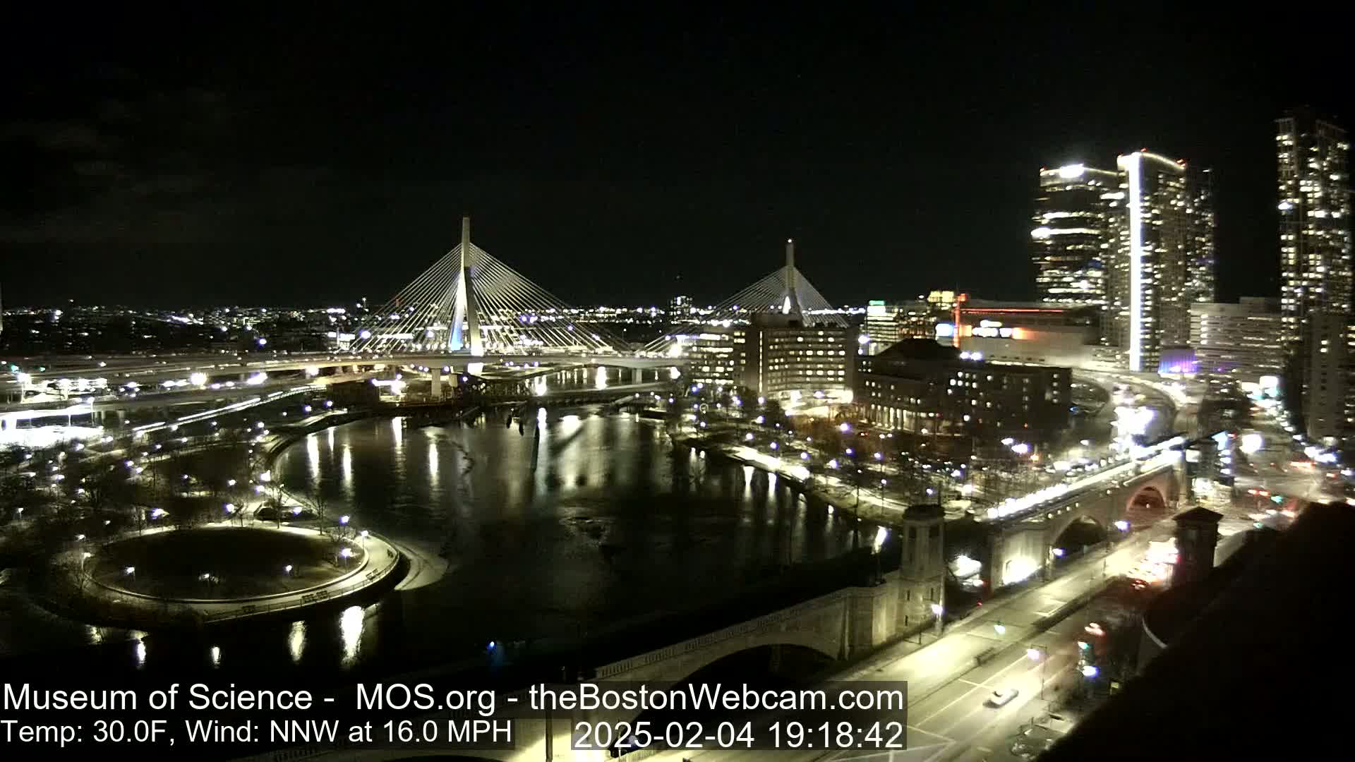 A nighttime cityscape featuring illuminated bridges spanning a river, city buildings, and roadways, with a 30°F temperature and a 16 mph northwest wind.