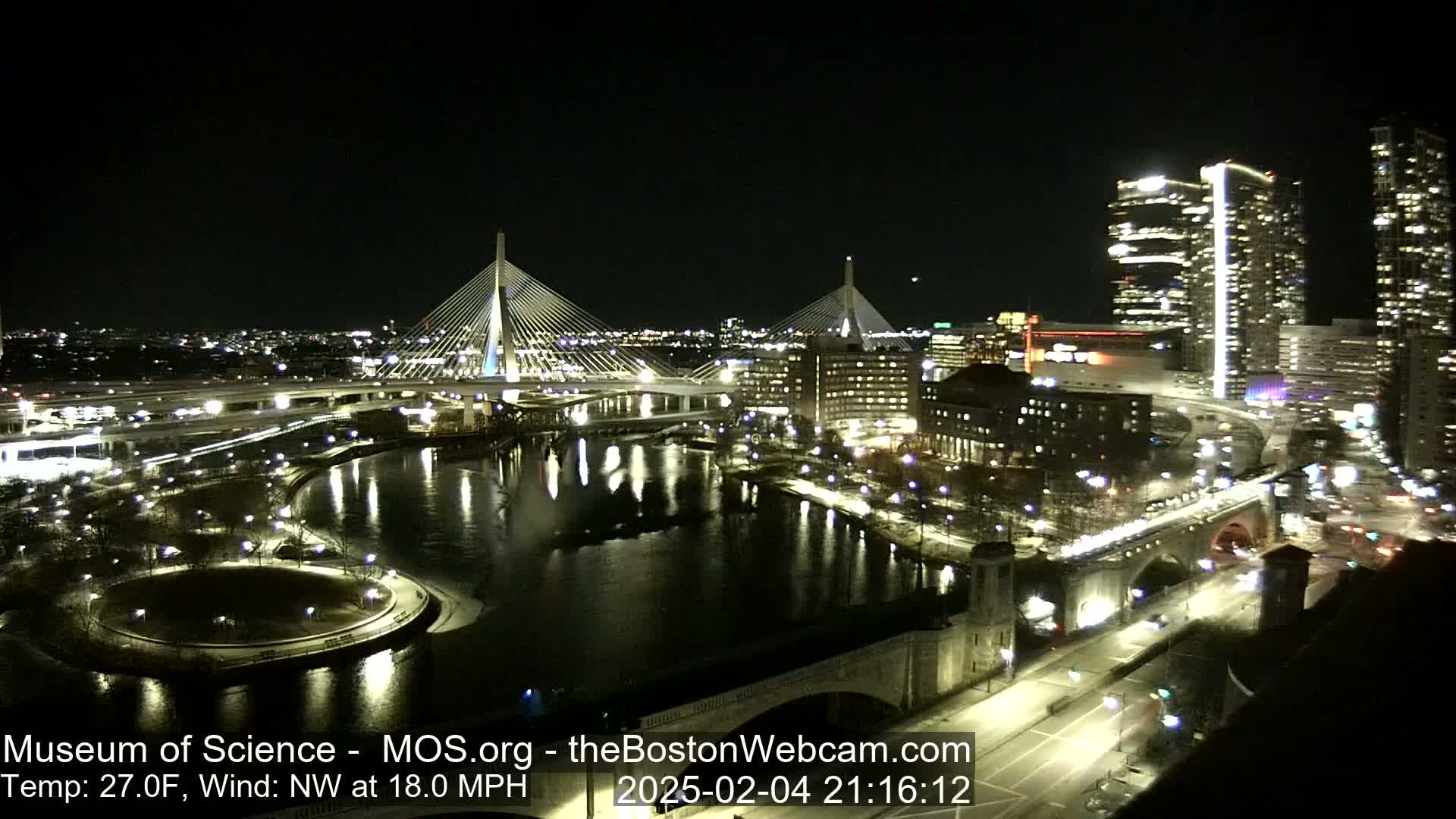 A nighttime cityscape with two cable-stayed bridges illuminated over a dark river, under a clear sky with a northwest wind at 18 mph and a temperature of 27°F.