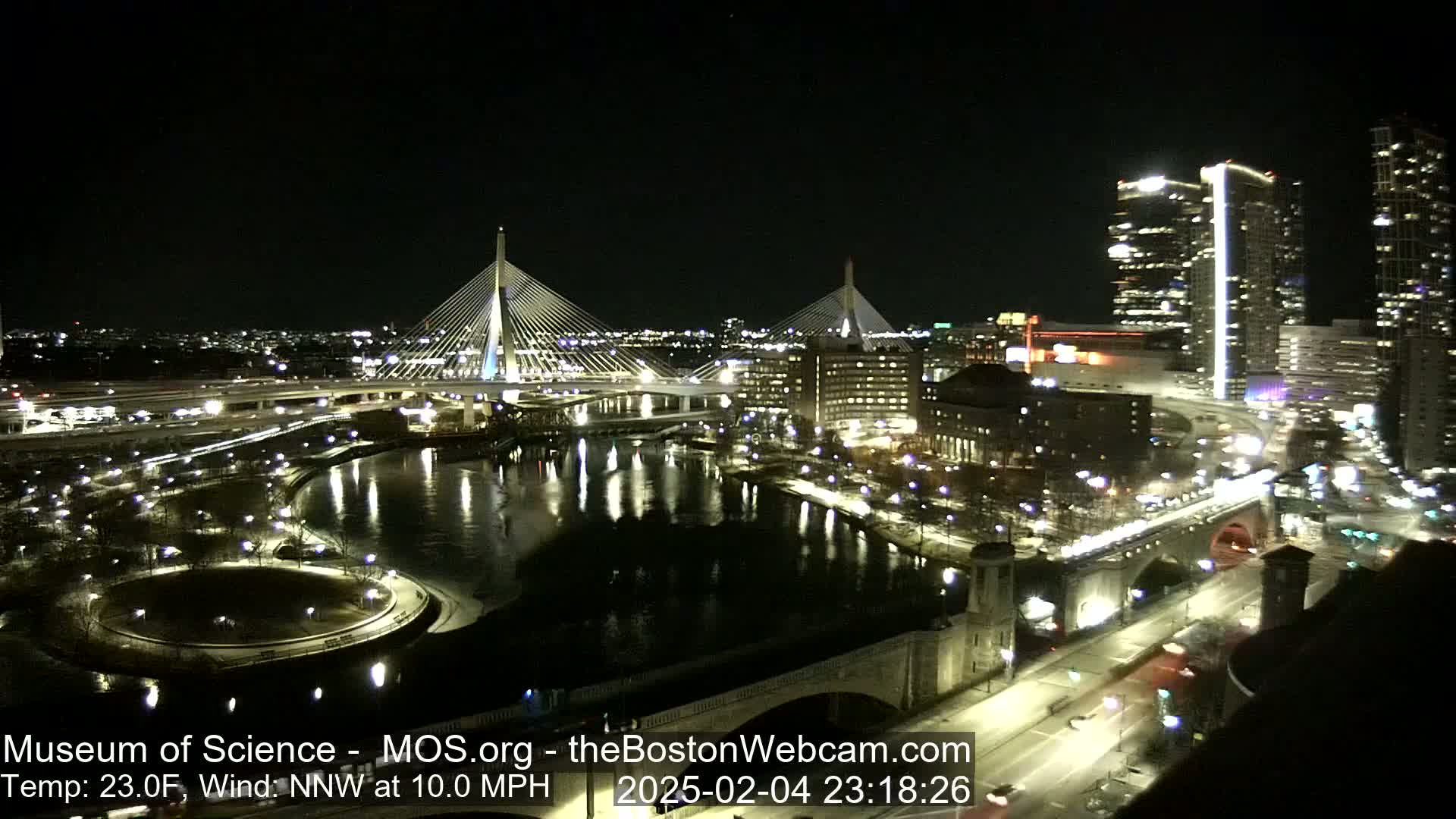 A nighttime cityscape with two cable-stayed bridges spanning a dark river, under a clear sky with a 23°F temperature and a 10 mph north-northwest wind.