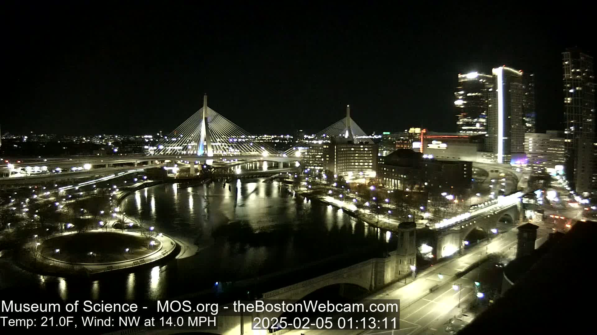 A nighttime, high-angle view of two illuminated cable-stayed bridges spanning a dark river, with city lights and buildings reflecting in the water, under a clear sky and a northwest wind at 14 mph.