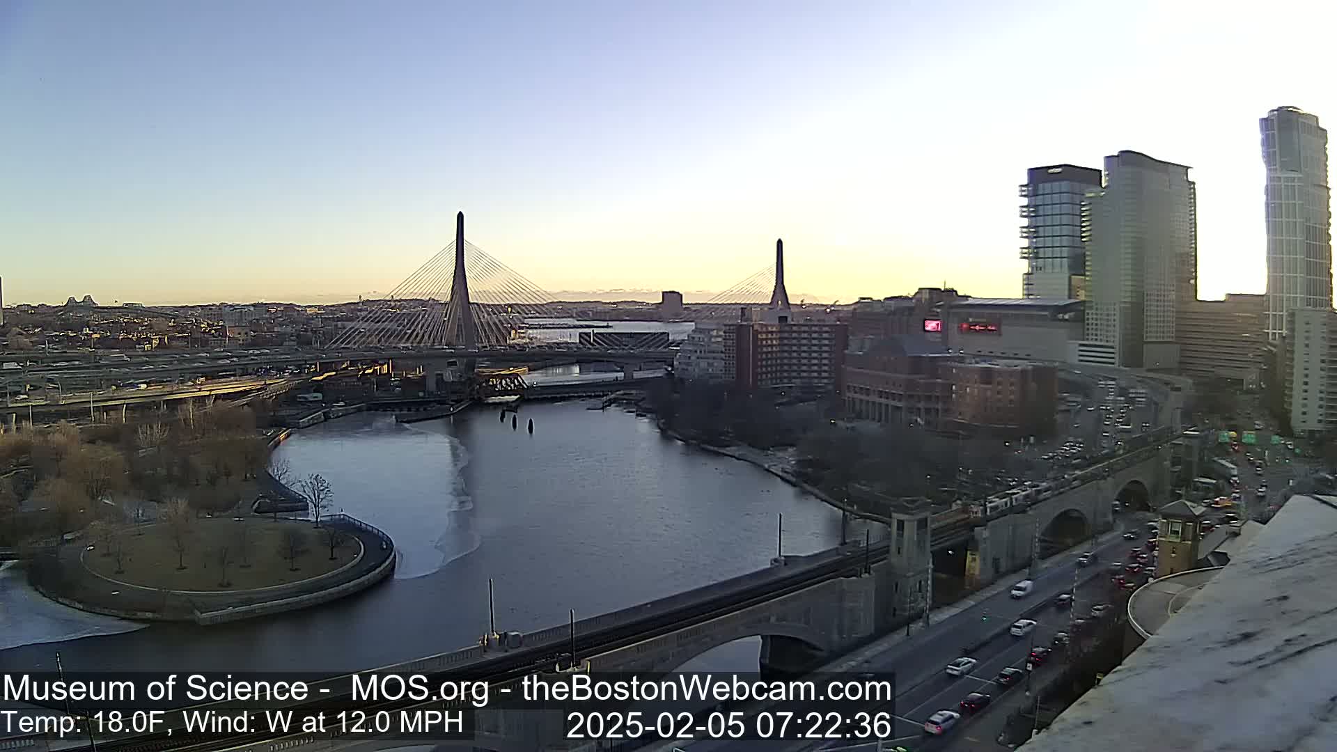 A cityscape at sunrise with a partially frozen river, two cable-stayed bridges, and city buildings, under a clear sky and a westerly wind of 12 mph at 18°F.