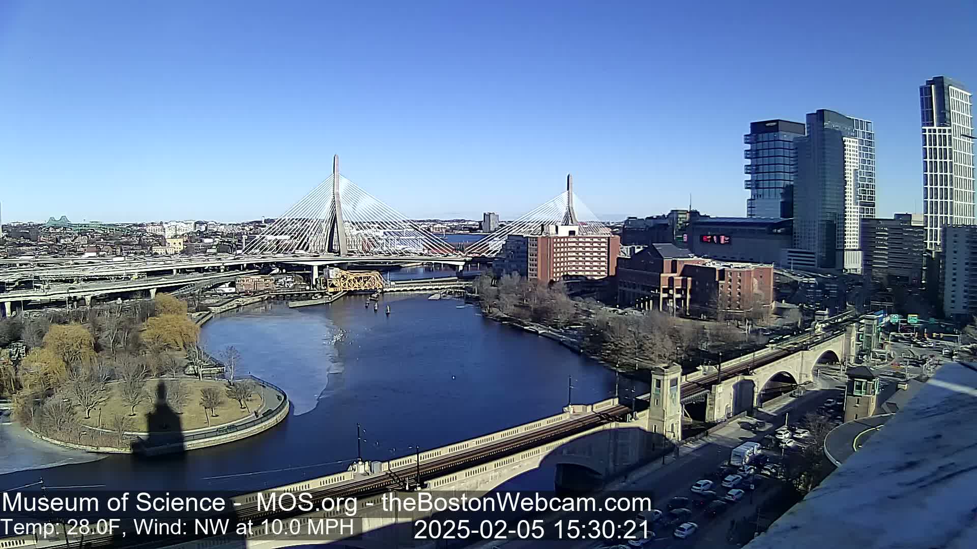 A sunny day reveals a cityscape with two cable-stayed bridges spanning a river, surrounded by buildings, roadways, and a park.