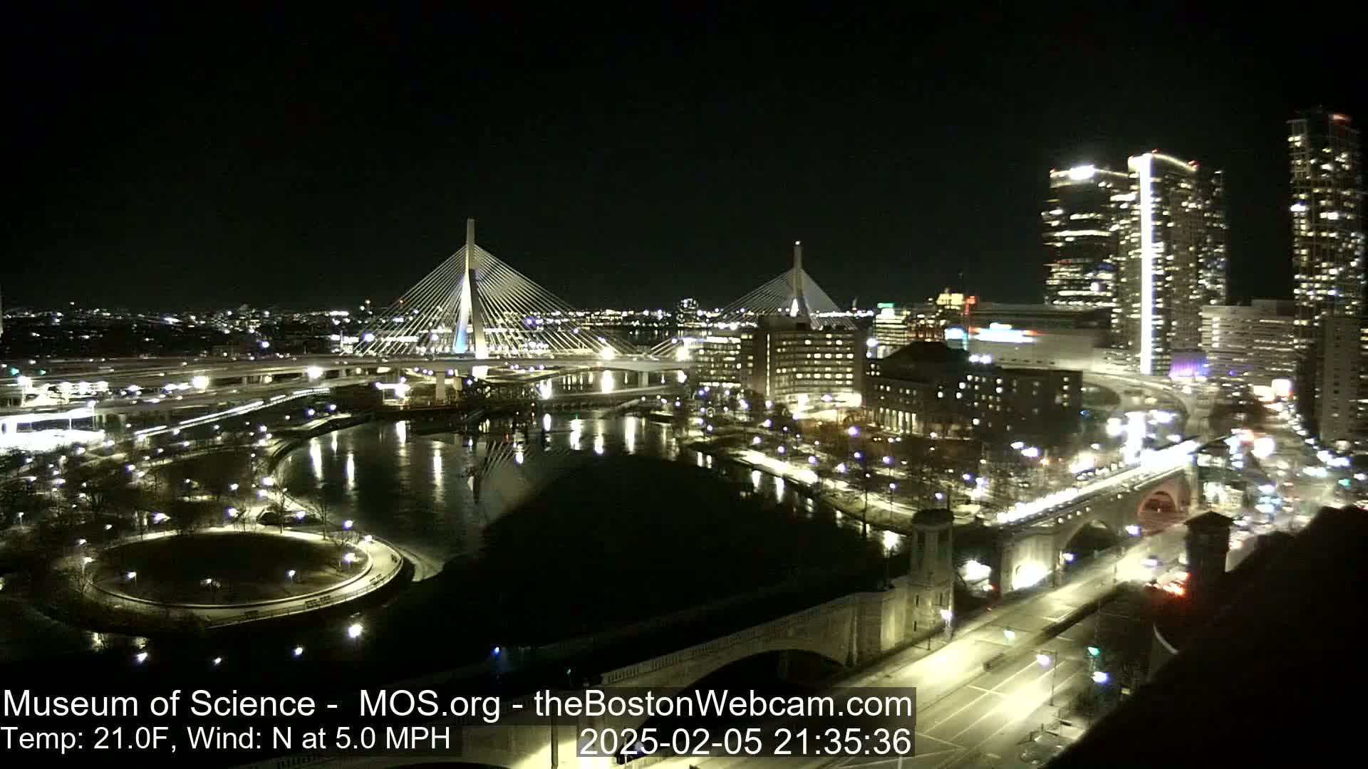 A nighttime city scene shows two cable-stayed bridges spanning a dark river, with city lights illuminating buildings and roadways under a clear sky.