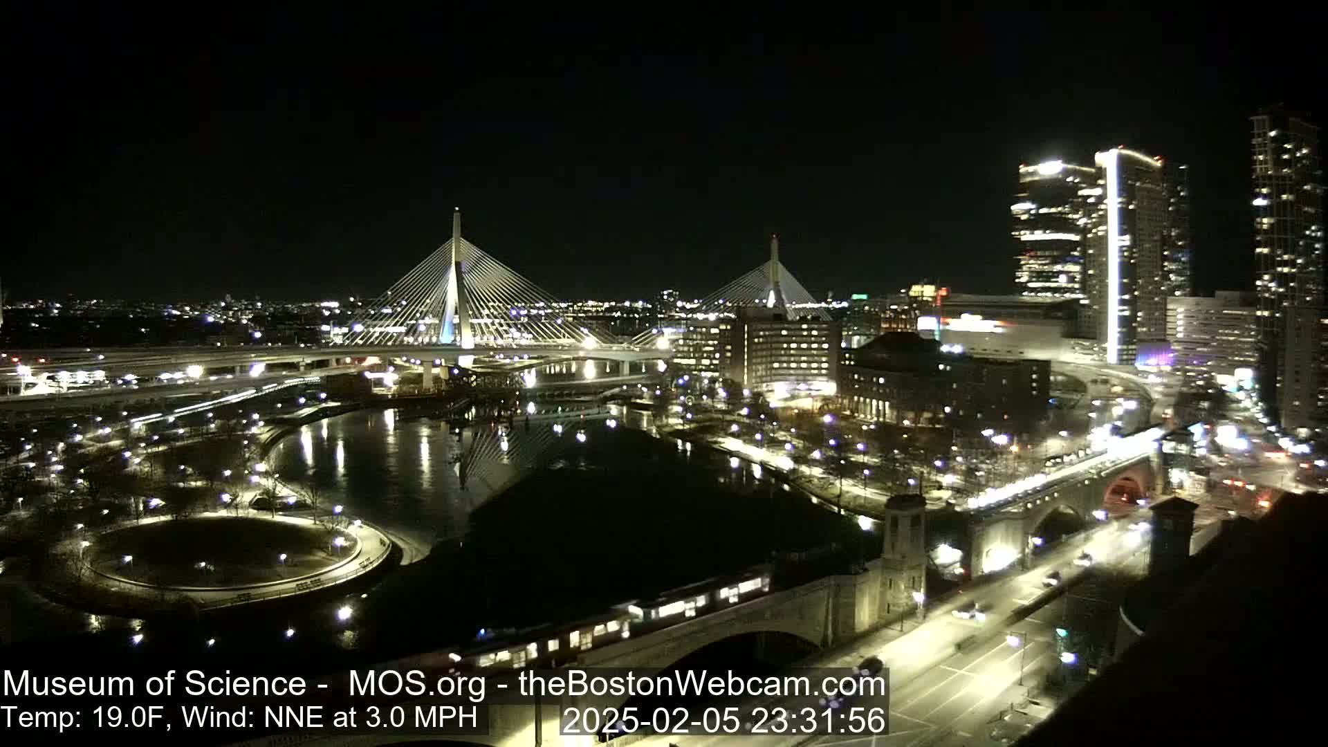 A nighttime cityscape shows two illuminated cable-stayed bridges spanning a dark river, with city lights reflecting on the water under a clear sky.