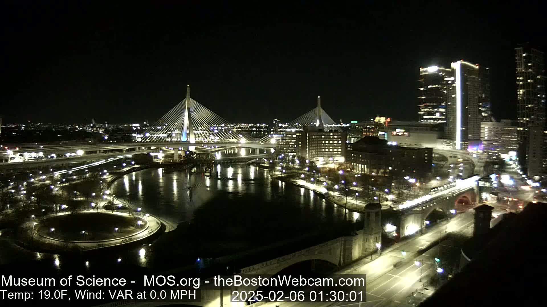 A nighttime view of two illuminated cable-stayed bridges spanning a dark river, with city lights and buildings reflecting in the water.
