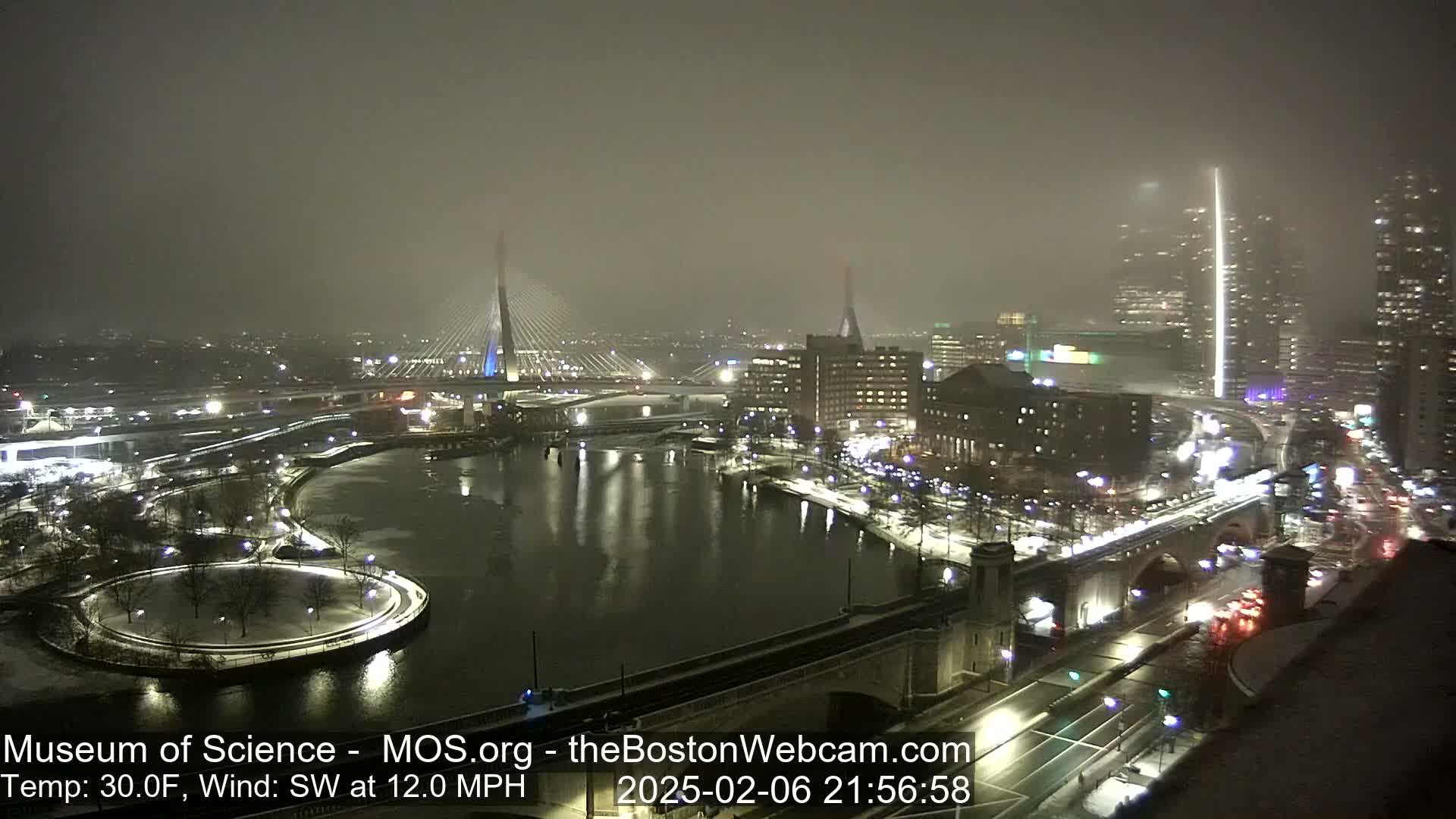 A nighttime, snowy scene of a city waterfront with two cable-stayed bridges, illuminated buildings, and roadways covered in snow, under a hazy, dark sky.
