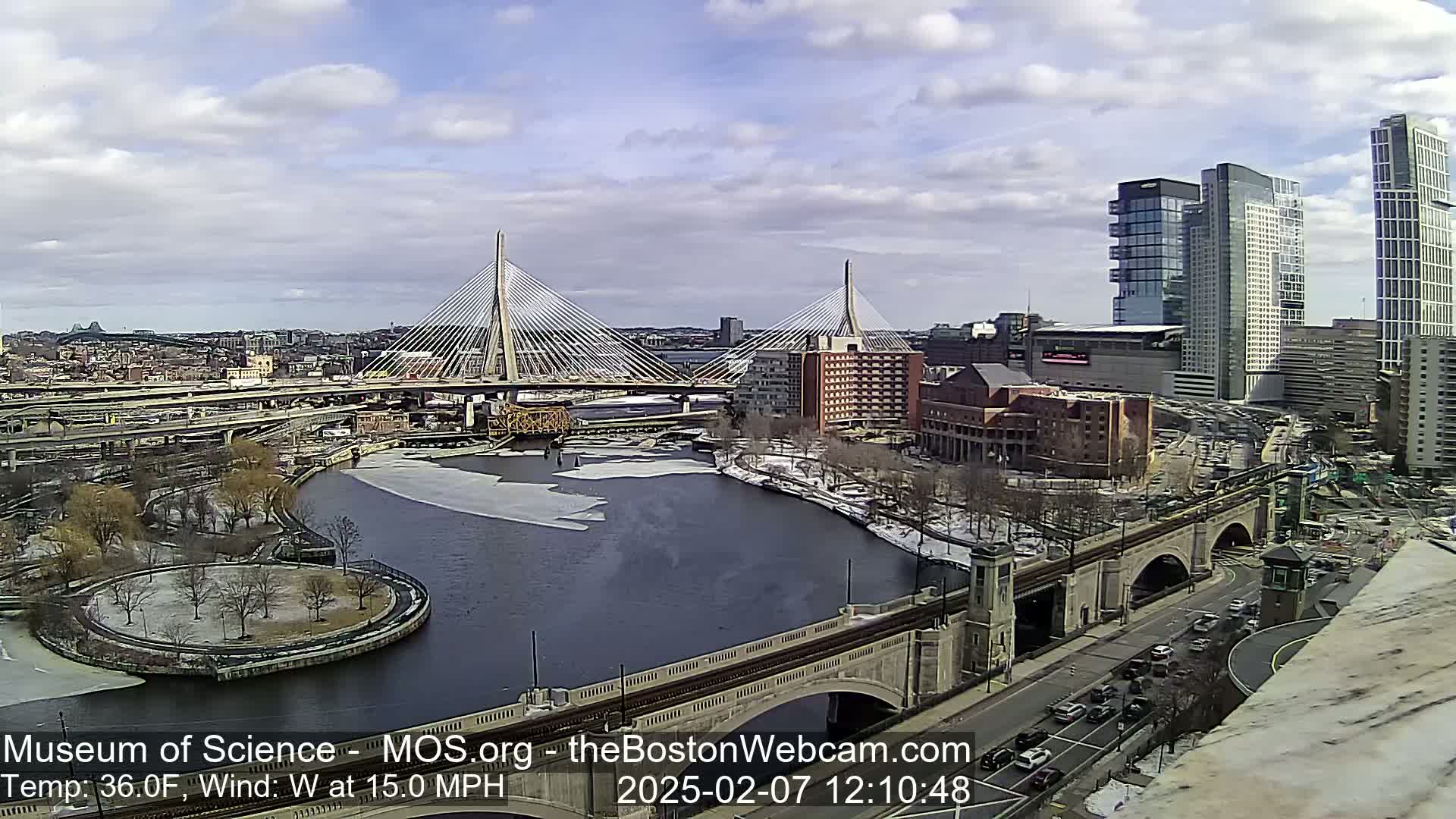 A partly cloudy day shows a city with two cable-stayed bridges spanning a partially frozen river, with a third bridge visible in the foreground.