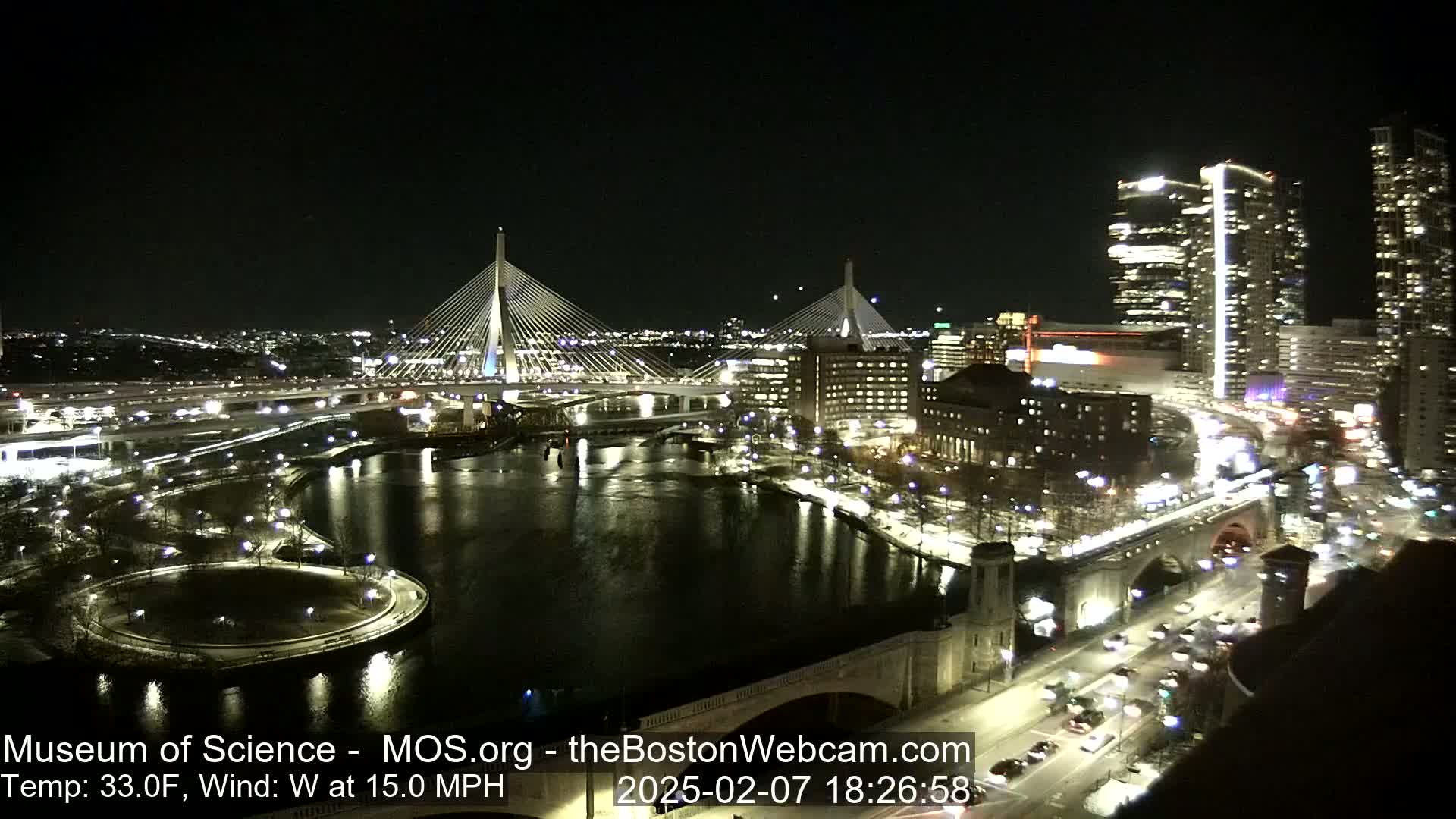 A nighttime cityscape shows two illuminated cable-stay bridges spanning a dark river, with city lights reflecting on the water under a clear sky, and a 15 mph west wind.