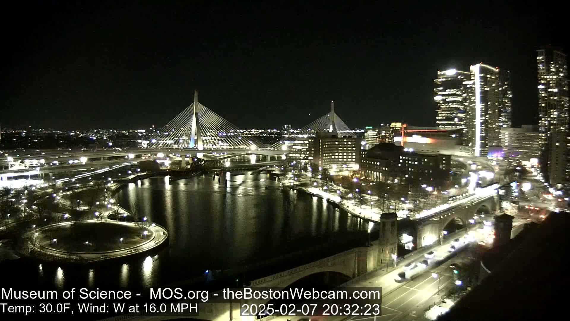 A nighttime view of a city with two cable-stayed bridges spanning a river, under a clear, dark sky with a temperature of 30°F and a 16 mph westerly wind.
