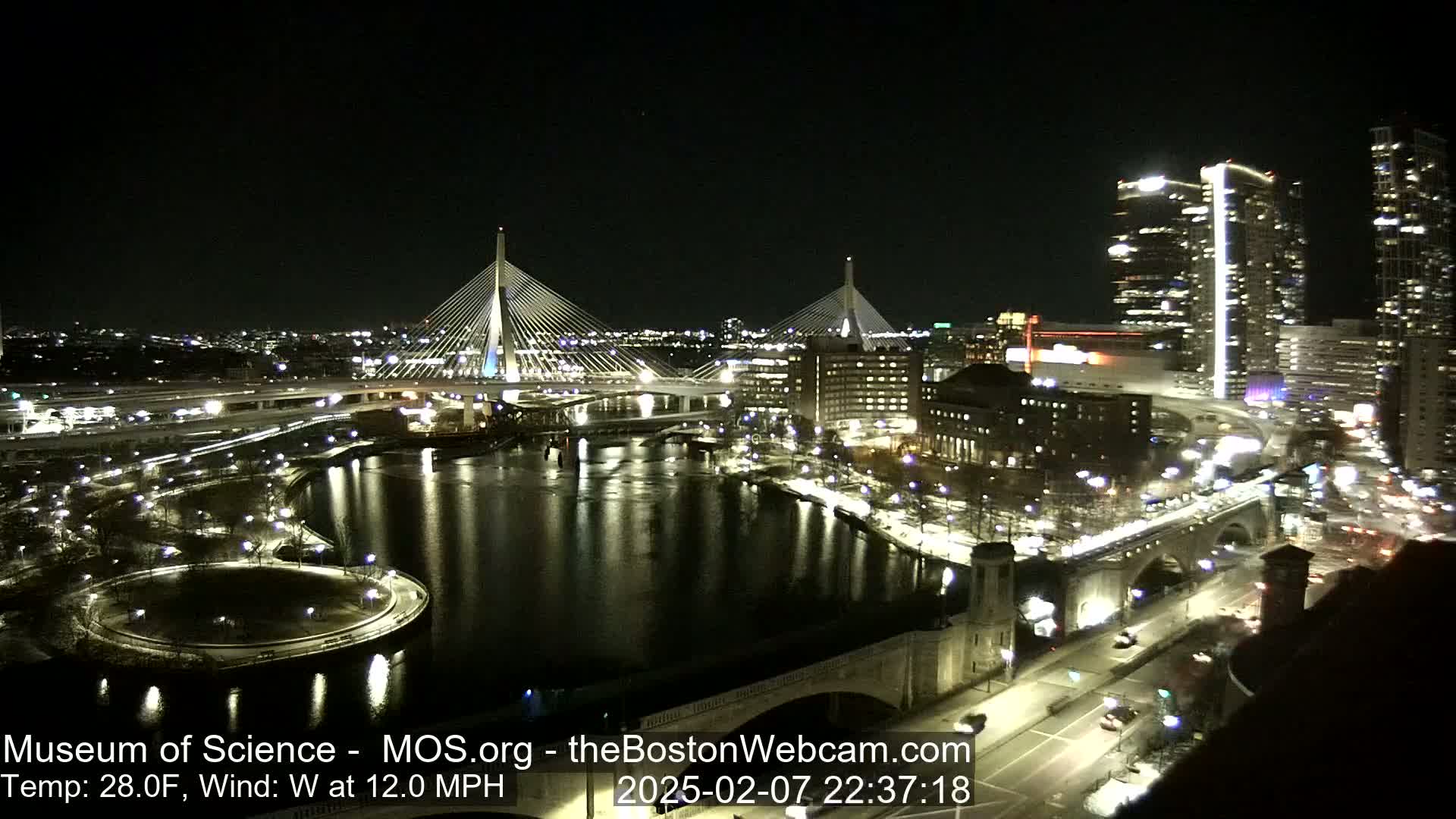 A nighttime view of a city waterfront, featuring two illuminated cable-stayed bridges, a river reflecting city lights, and roadways with sparse traffic under a clear sky with a temperature of 28 degrees Fahrenheit and a 12 mph west wind.