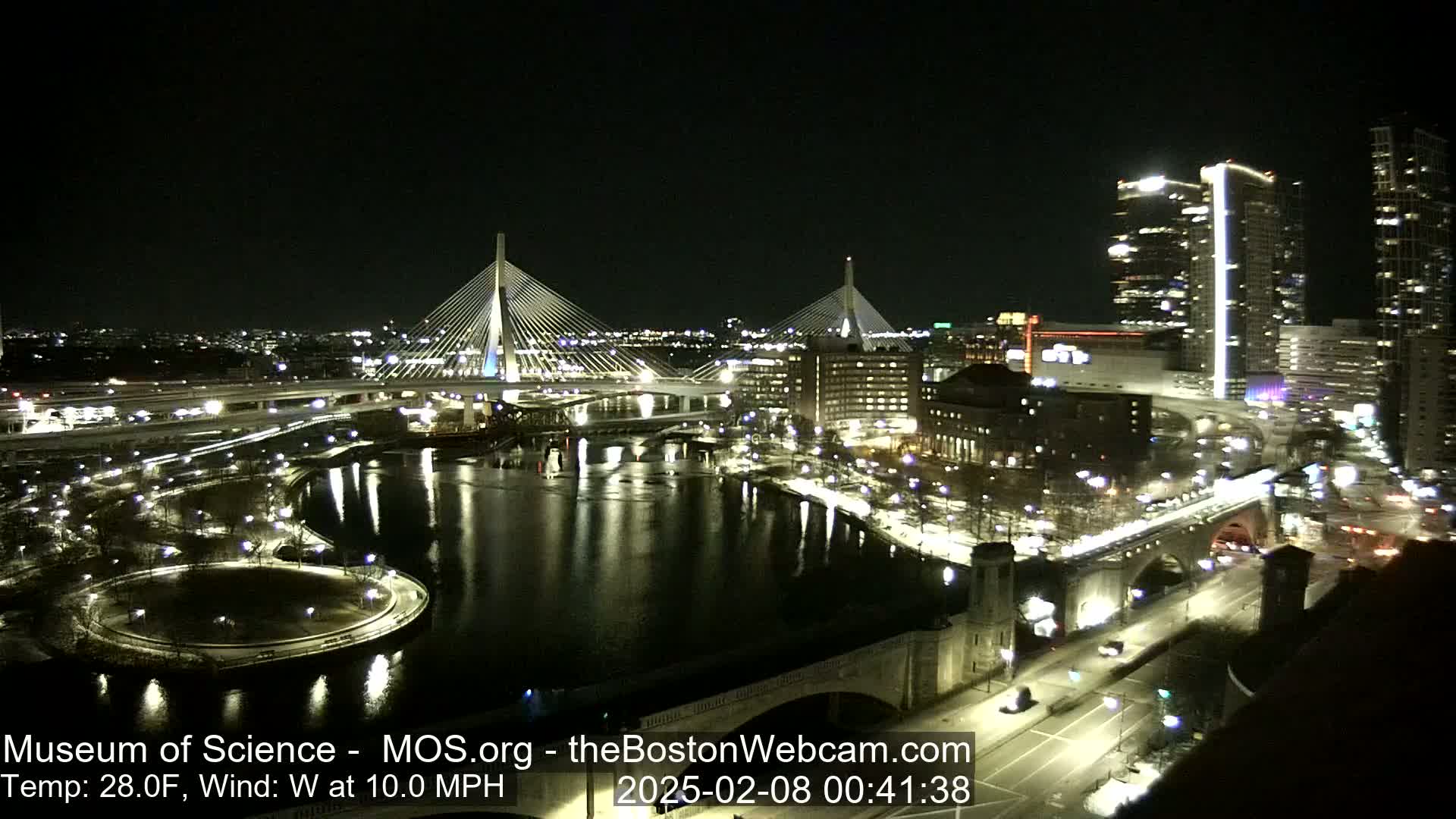 A nighttime view of a city with two illuminated cable-stayed bridges spanning a river, with a temperature of 28 degrees Fahrenheit and a 10 mph westerly wind.