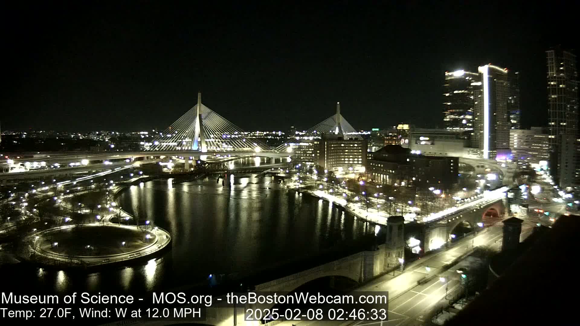 A nighttime view of a city illuminated with lights, featuring two cable-stayed bridges spanning a dark river, under a clear sky with a temperature of 27°F and a 12 mph westerly wind.