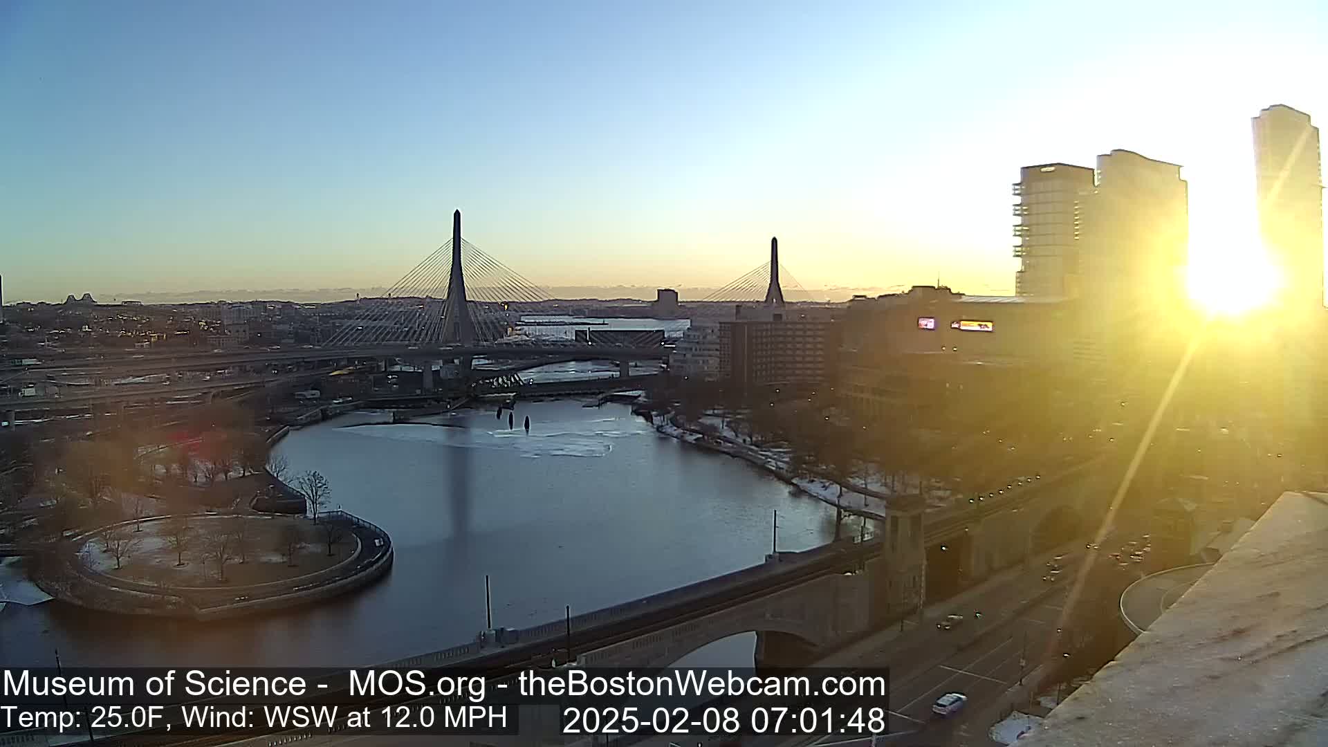 A sunrise casts a bright glow over a cityscape featuring two cable-stayed bridges spanning a partially frozen river, with a city skyline and buildings in the background under a clear sky.