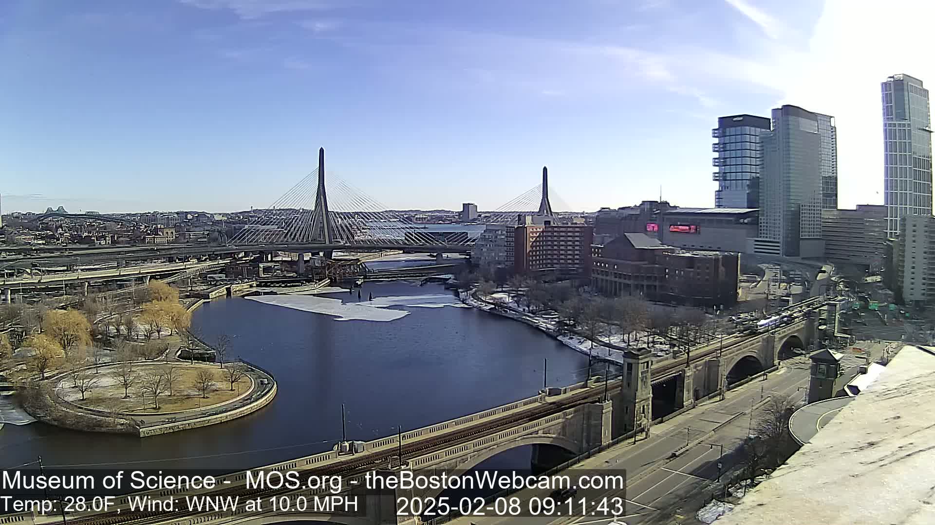 A sunny day reveals a cityscape with two cable-stayed bridges spanning a river partially covered in ice, alongside other bridges and high-rise buildings.