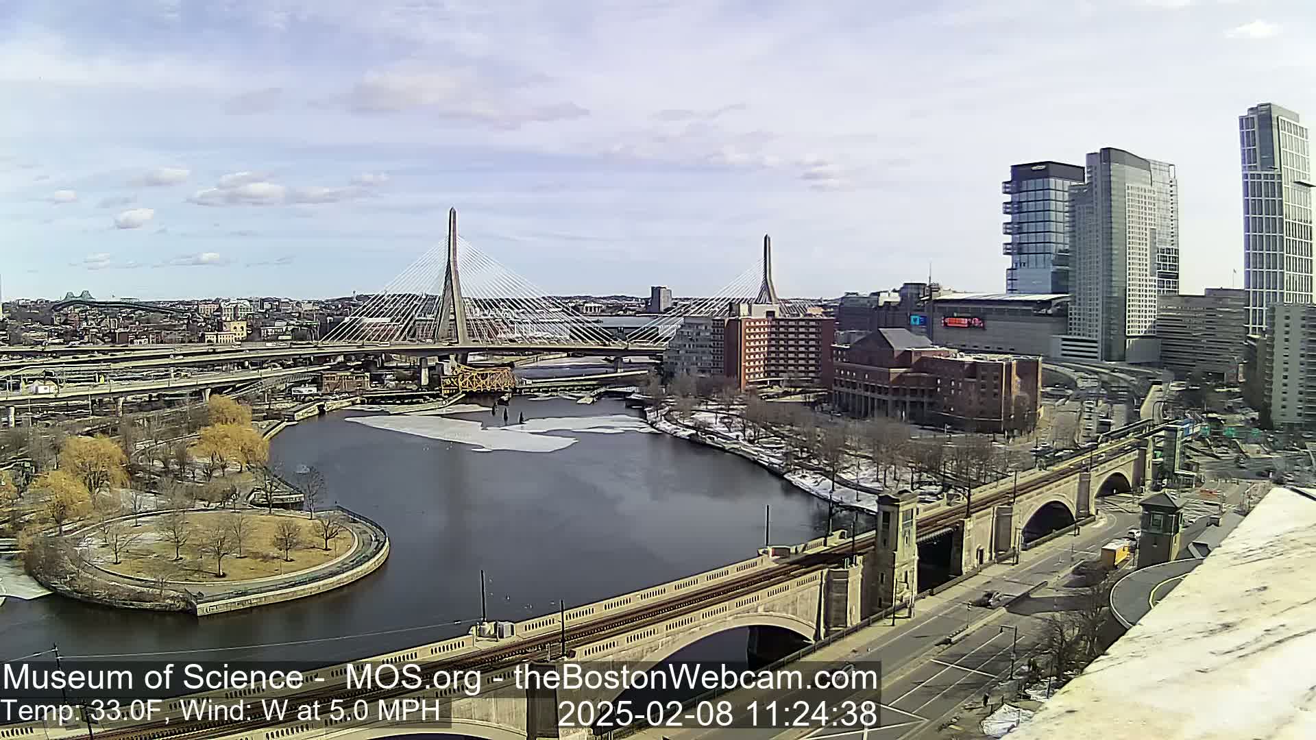 A partly sunny day reveals a cityscape with two cable-stayed bridges spanning a river partially covered in ice, alongside other bridges and modern high-rise buildings.