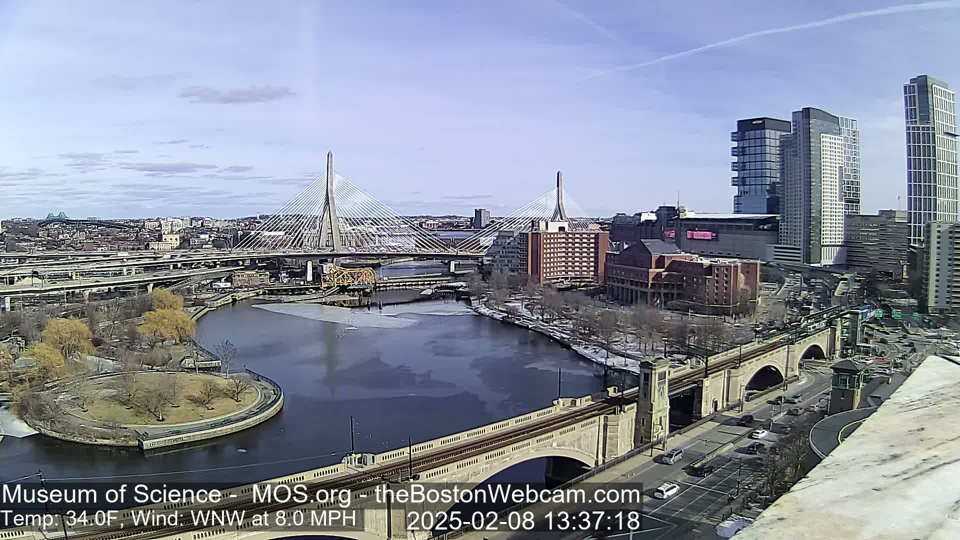 A sunny day reveals a cityscape with two cable-stayed bridges spanning a river partially covered in ice, alongside other bridges and modern high-rise buildings.