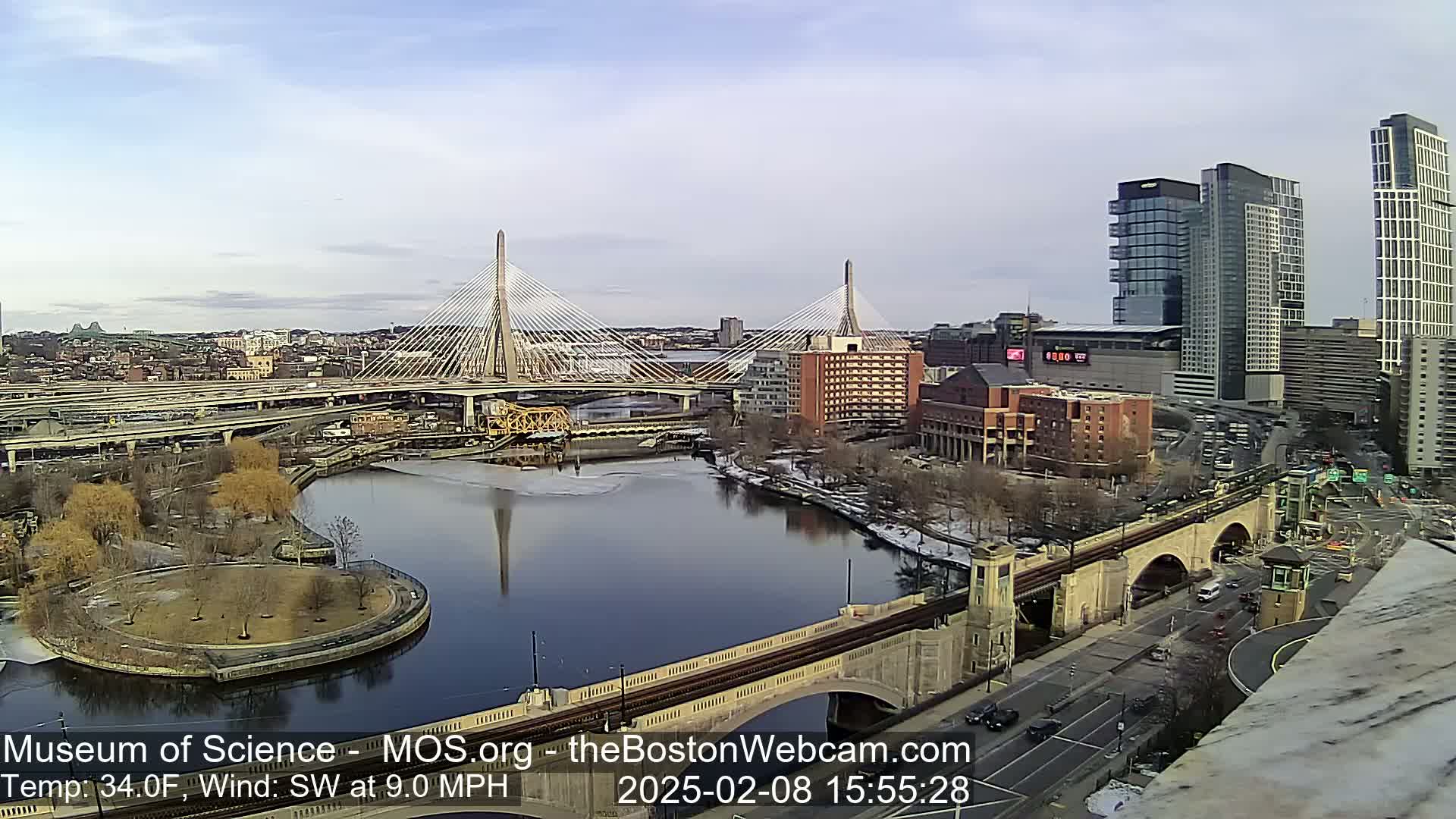 A partly sunny day reveals a cityscape with two cable-stayed bridges spanning a river, partially frozen, with modern and historic buildings, and a park.