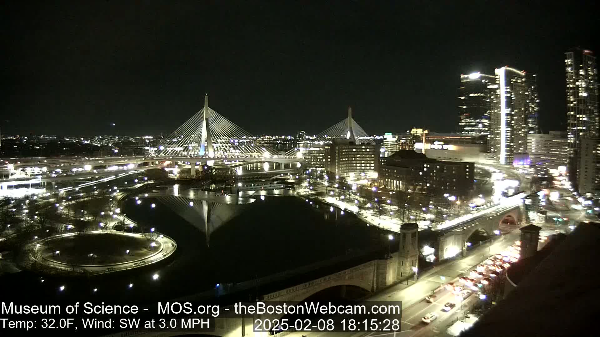 A nighttime aerial view of a city with two illuminated cable-stayed bridges spanning a dark body of water, under a clear sky with a temperature of 32 degrees Fahrenheit and a southwest wind.