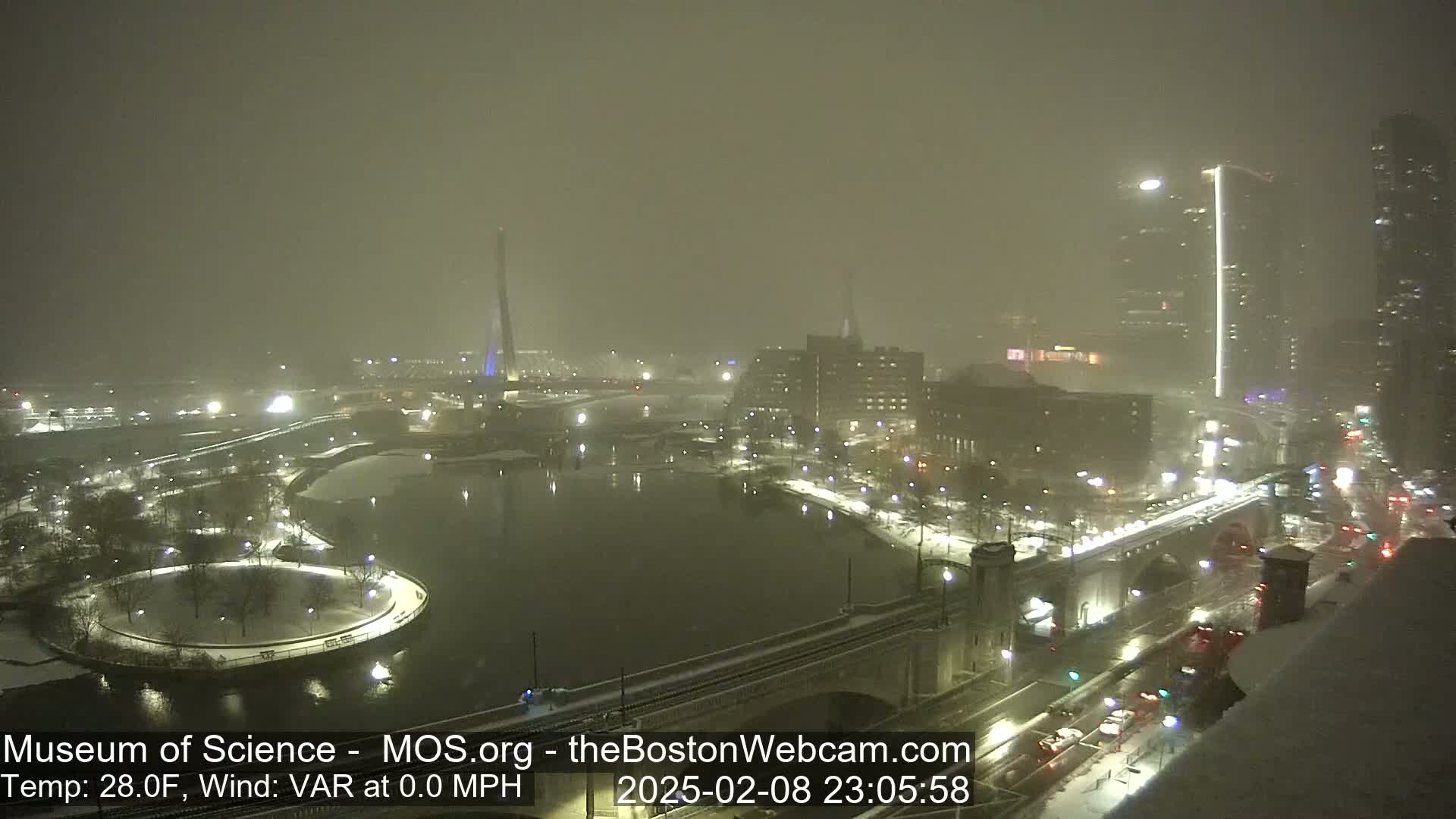 A snow-covered cityscape at night, viewed from a high vantage point overlooking a snow-covered bridge and a body of water.