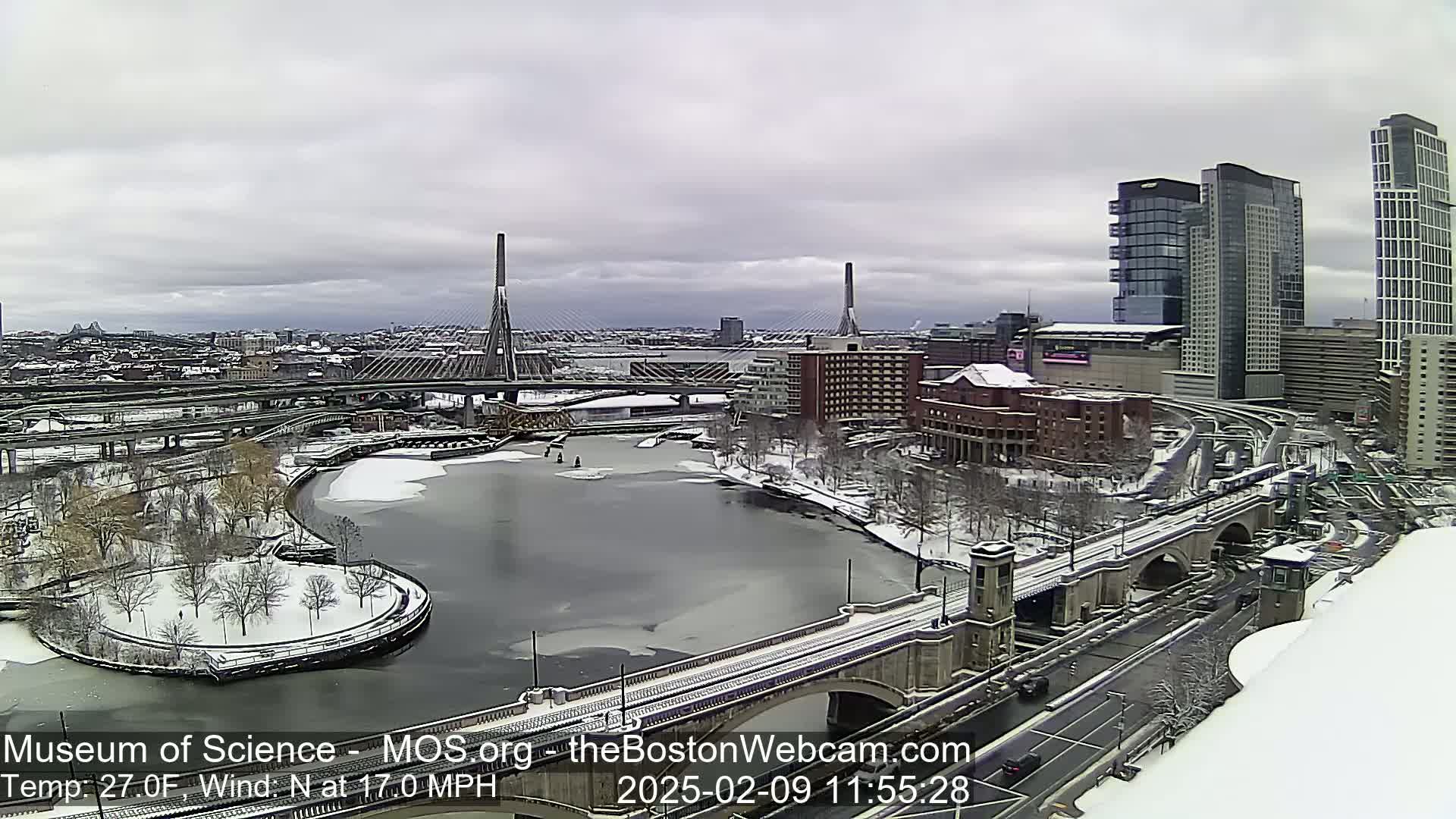 A snow-covered city landscape with a partially frozen river, bridges, and modern high-rises under a cloudy sky.