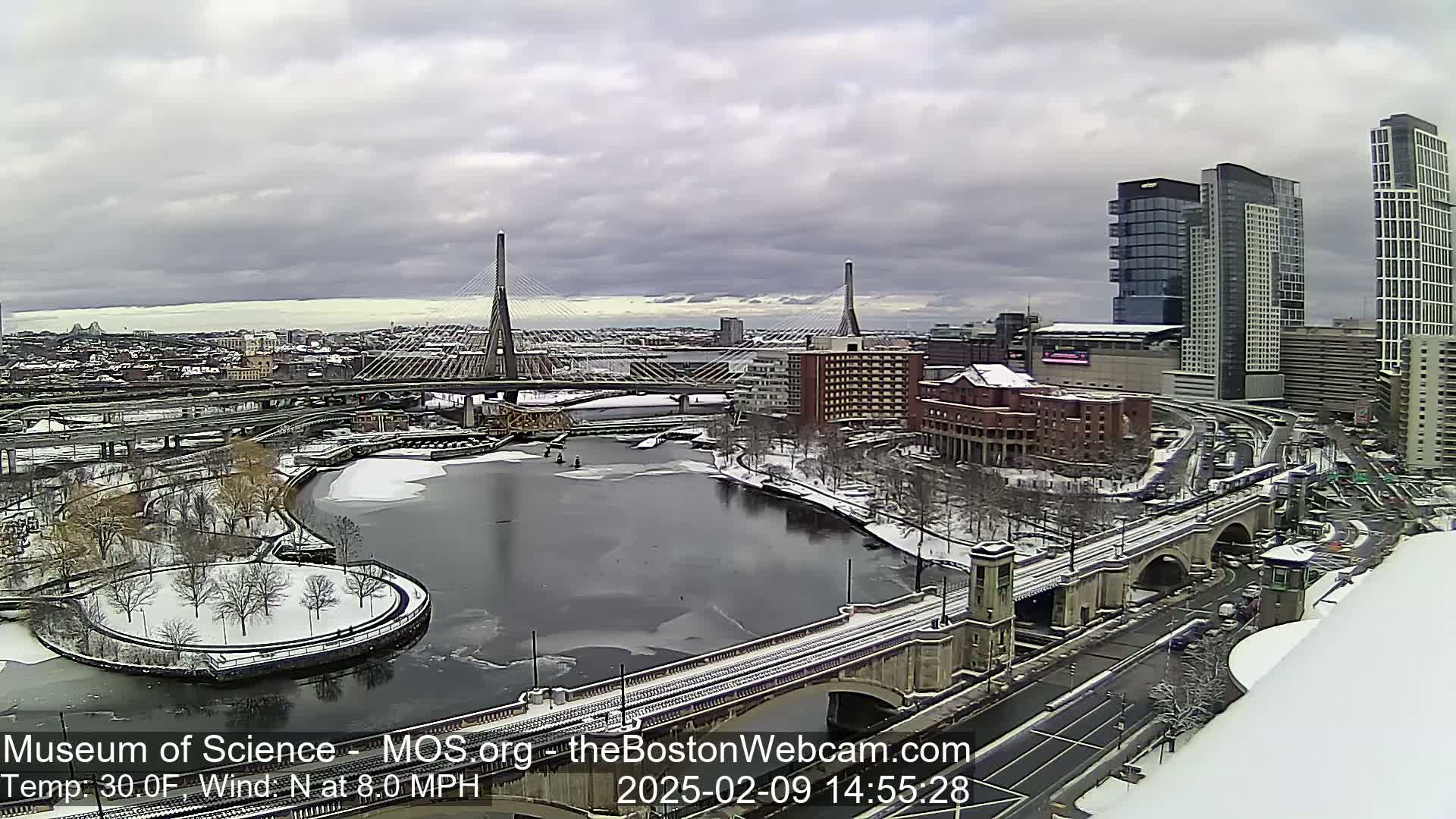 A snow-covered cityscape with a river partially frozen, featuring a cable-stayed bridge and high-rise buildings under an overcast sky.
