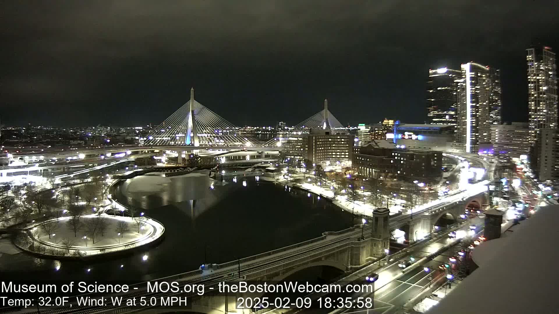 A nighttime view of a city with snow-covered ground, bridges illuminated, and a river reflecting city lights under a dark sky.
