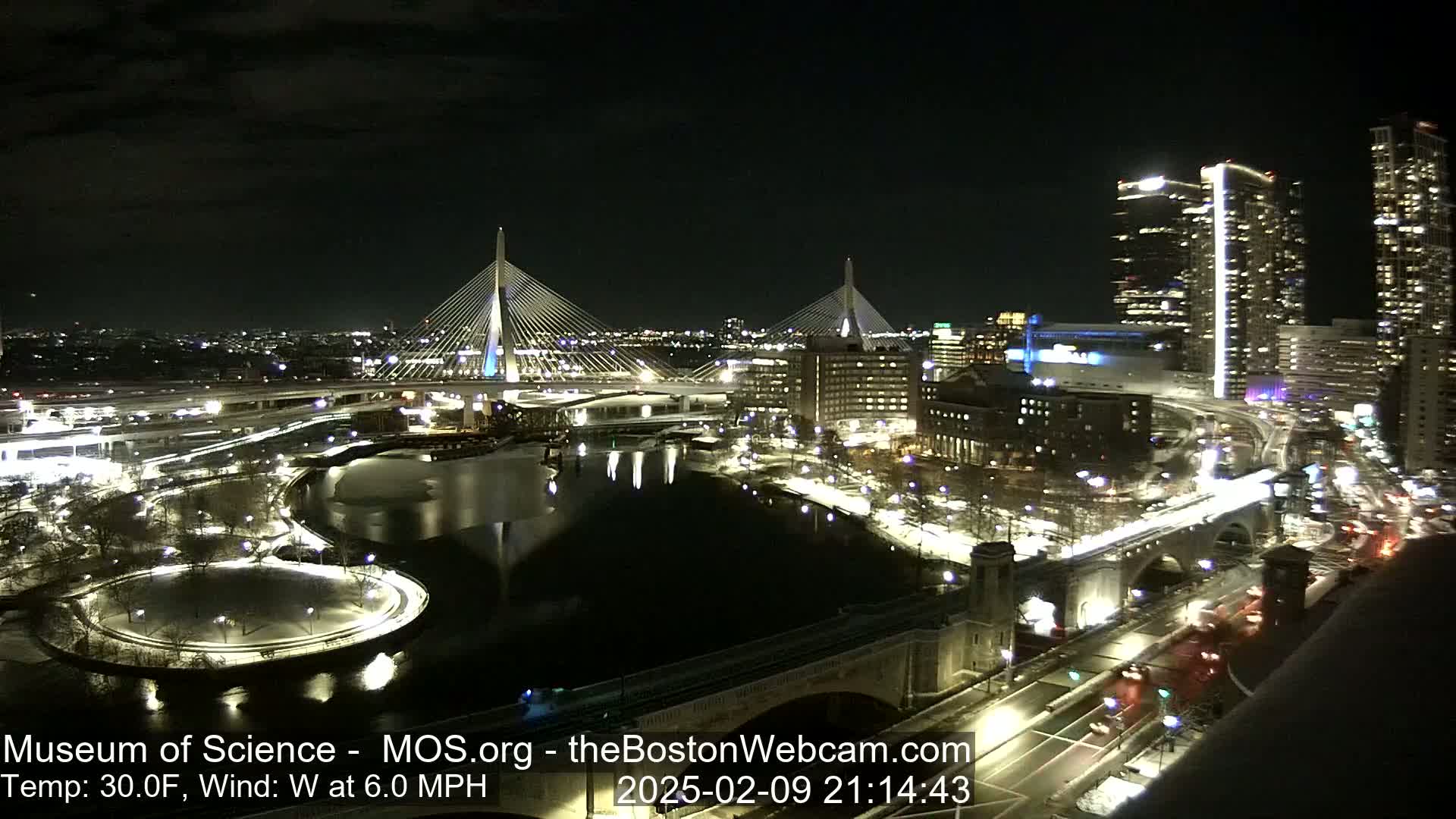 A nighttime view of a city with two illuminated cable-stayed bridges spanning a dark river, surrounded by city lights and snow-covered ground, with a west wind at 6.0 mph and a temperature of 30°F.
