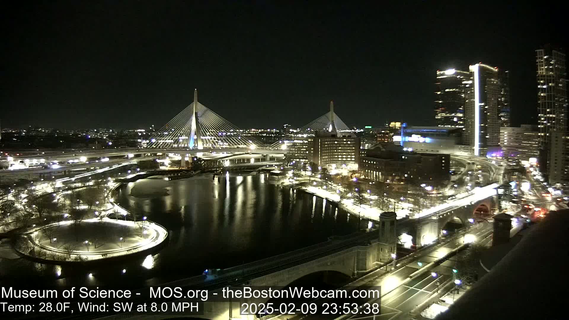 A nighttime cityscape with two cable-stayed bridges spanning a river, partially frozen, under a clear, dark sky with a southwest wind.