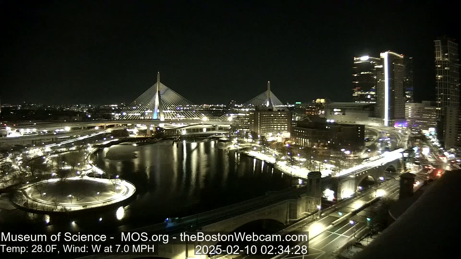 A nighttime, snow-covered cityscape with two cable-stayed bridges illuminated over a calm river, under a clear sky with a temperature of 28°F and a 7.0 mph westerly wind.