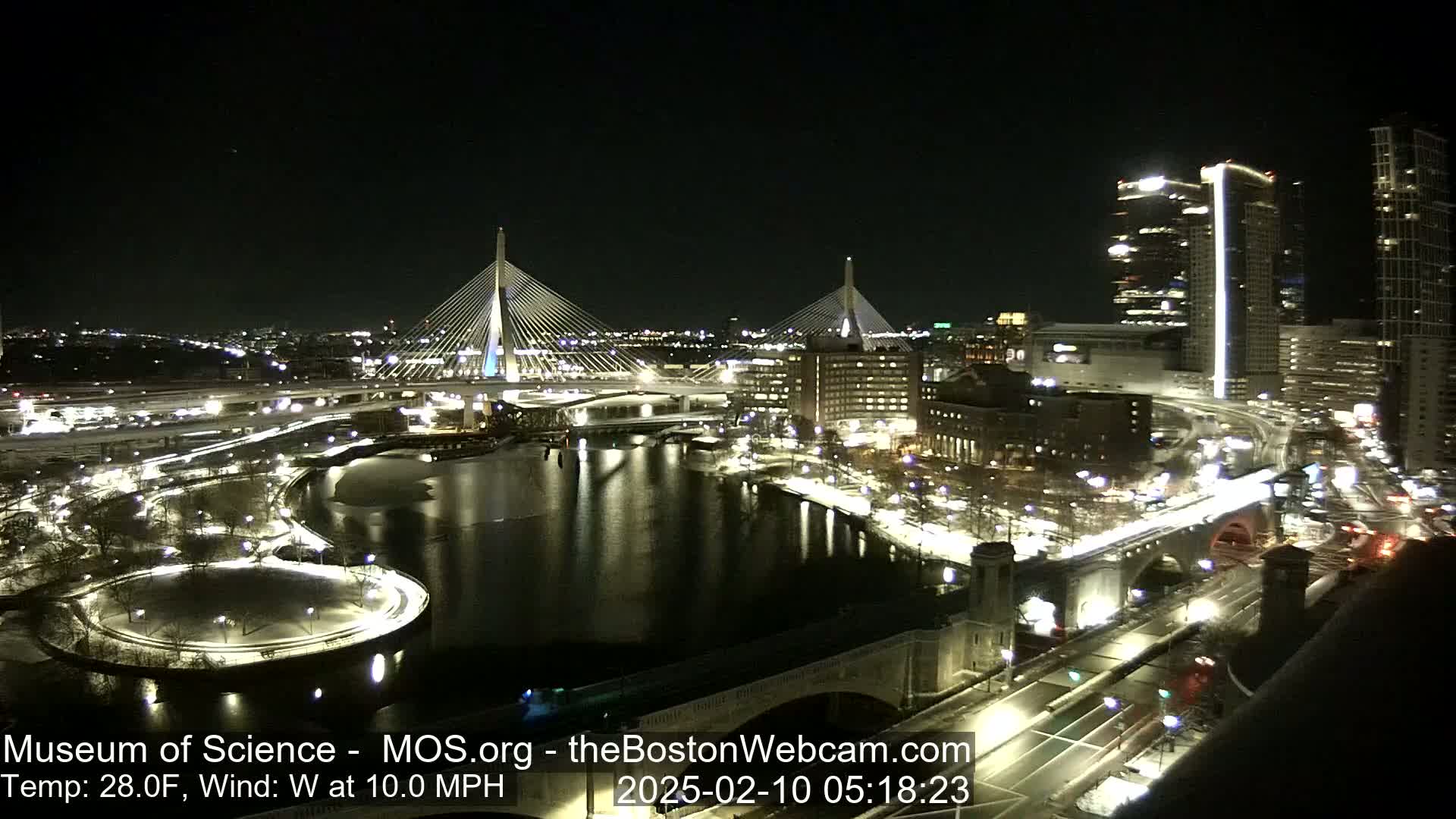 A nighttime cityscape with two cable-stayed bridges spanning a river, illuminated roadways, and buildings, under a clear, cold sky with a west wind.
