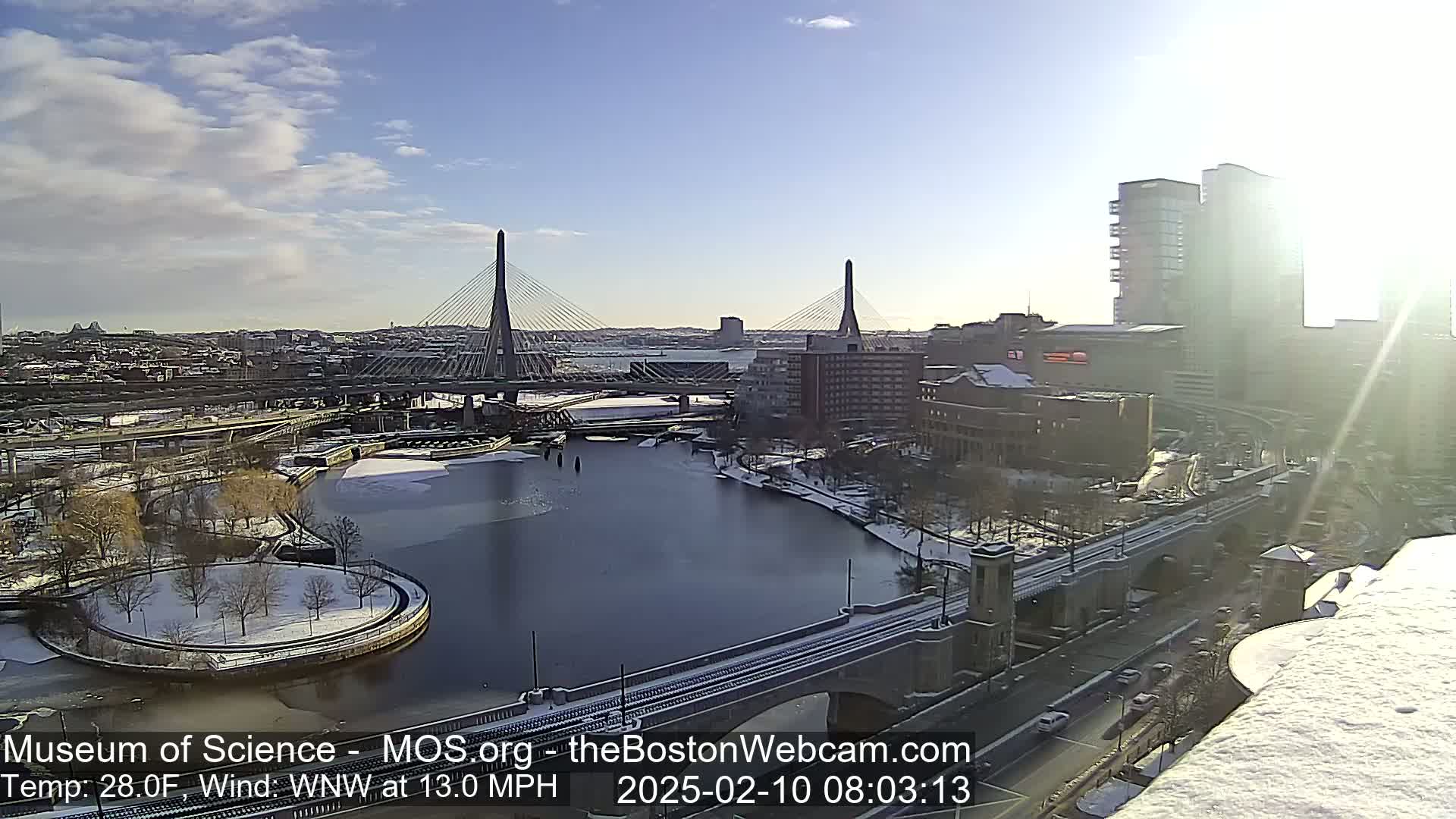 A sunny, cold day with a partly cloudy sky shows a cityscape with two cable-stayed bridges spanning a partially frozen river, surrounded by snow-covered parkland and buildings.